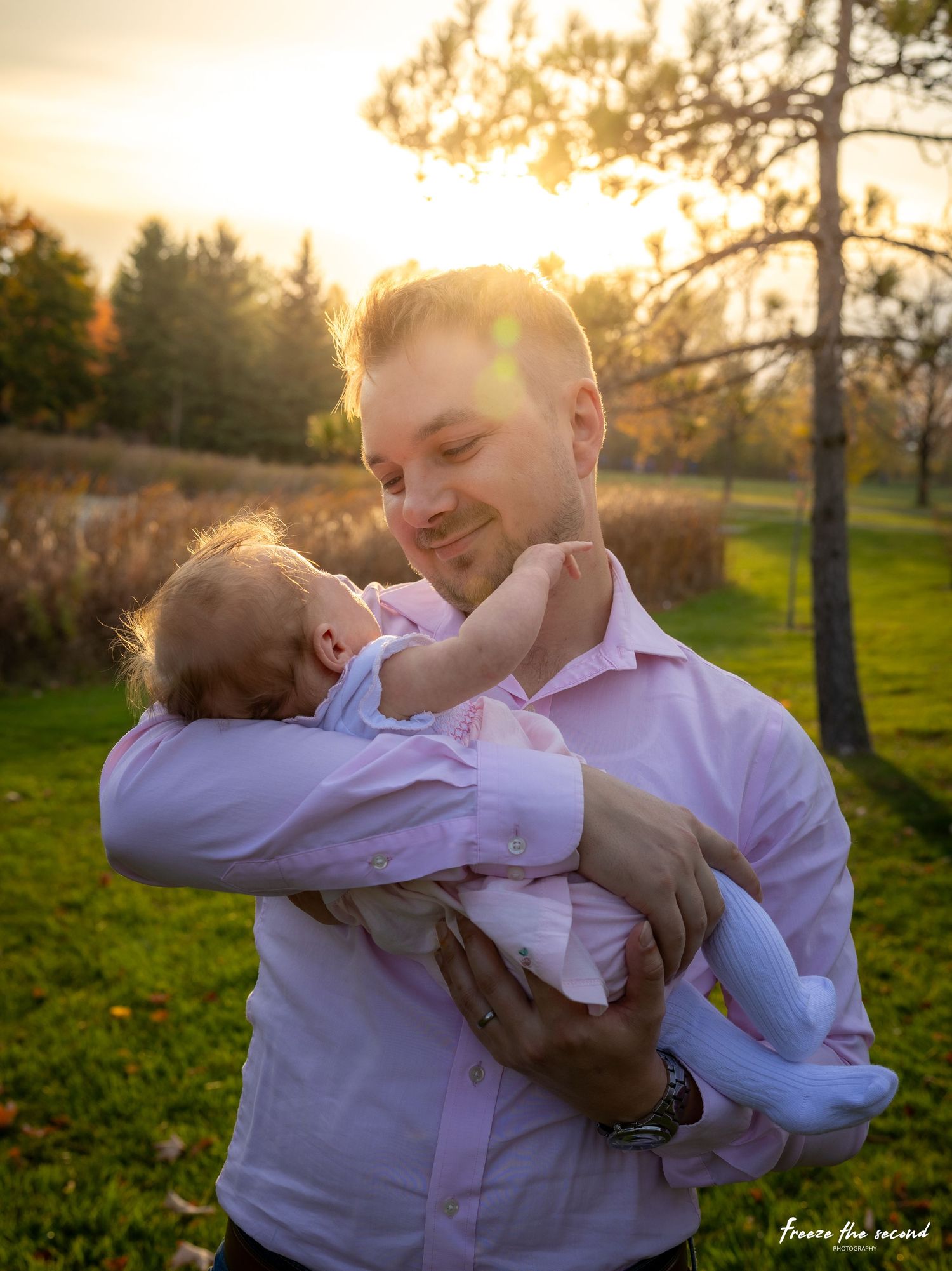 Father in pink shirt tenderly holds newborn baby at golden sunset in park setting.