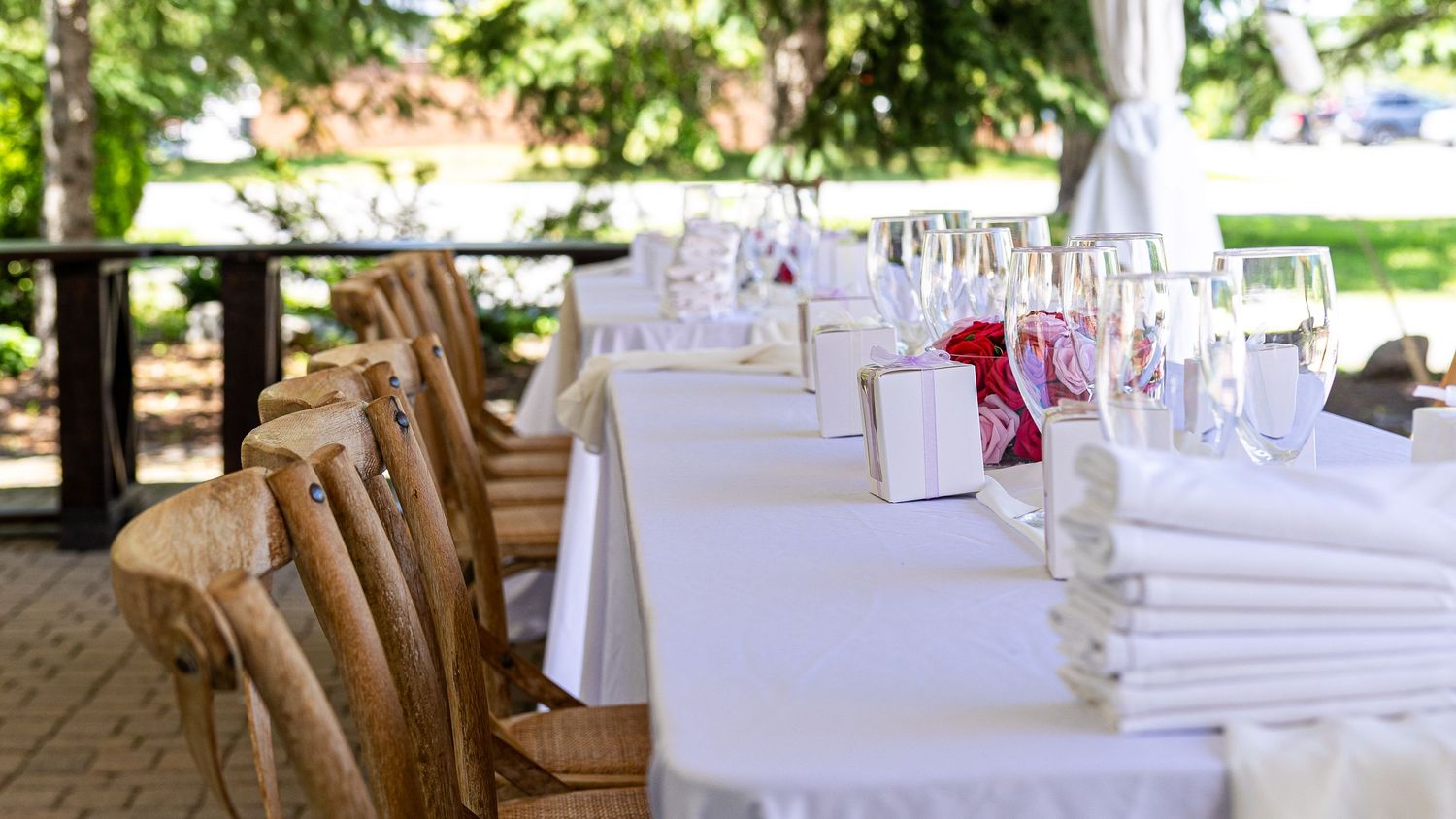 Outdoor wedding reception tables with white linens, wooden chairs, and glassware set in a sunny garden setting.