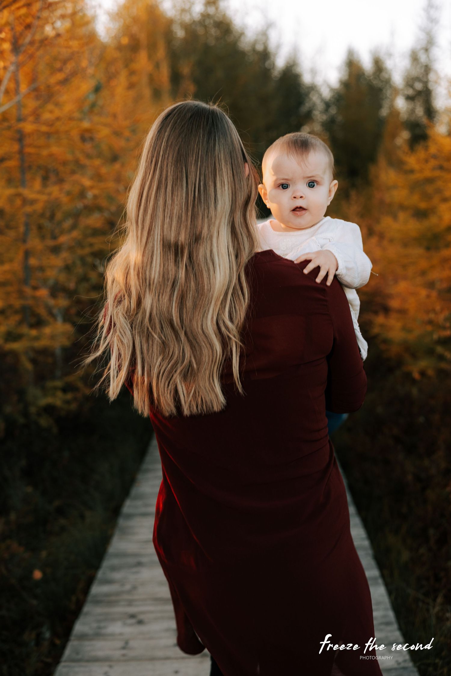Mother in burgundy dress holds baby on autumn boardwalk trail surrounded by golden foliage.
