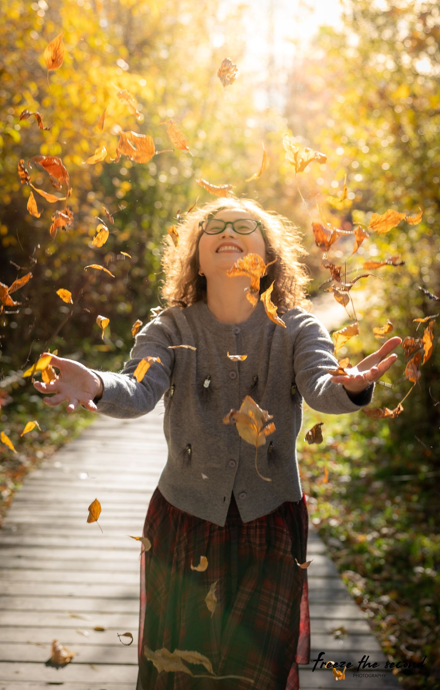 A smiling woman tosses autumn leaves in the air on a sunny fall boardwalk.