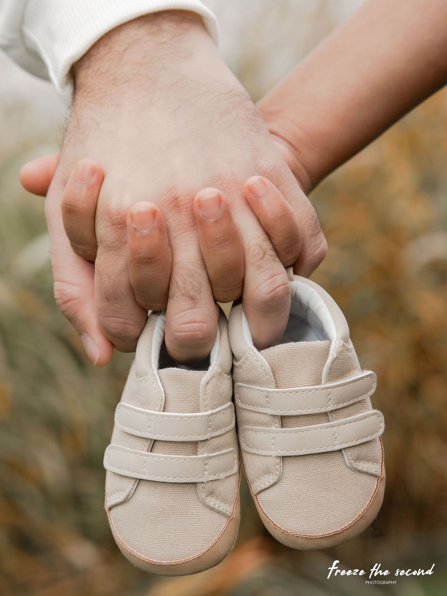 Two hands holding tiny baby sneakers outdoors, announcing pregnancy with cute neutral-toned infant shoes.