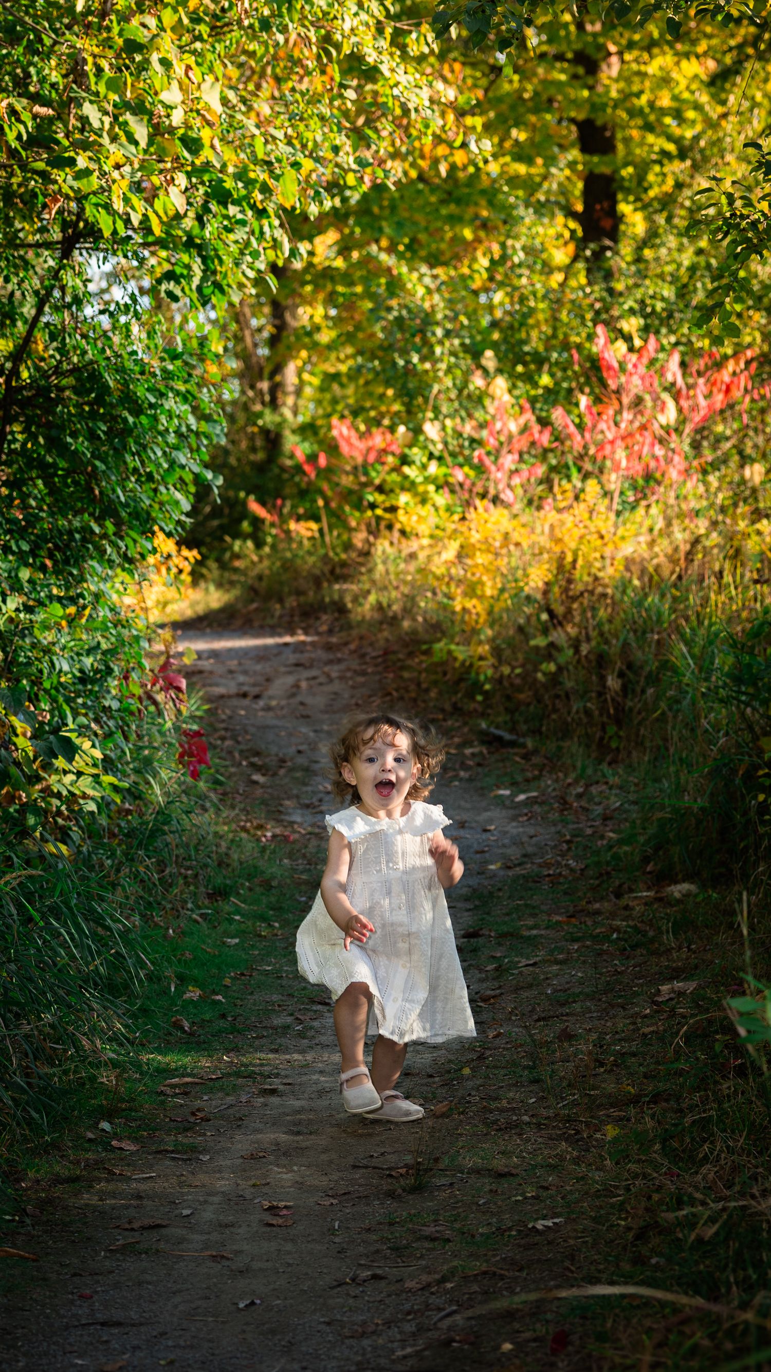 A joyful toddler in a white dress runs down a colorful autumn forest path.