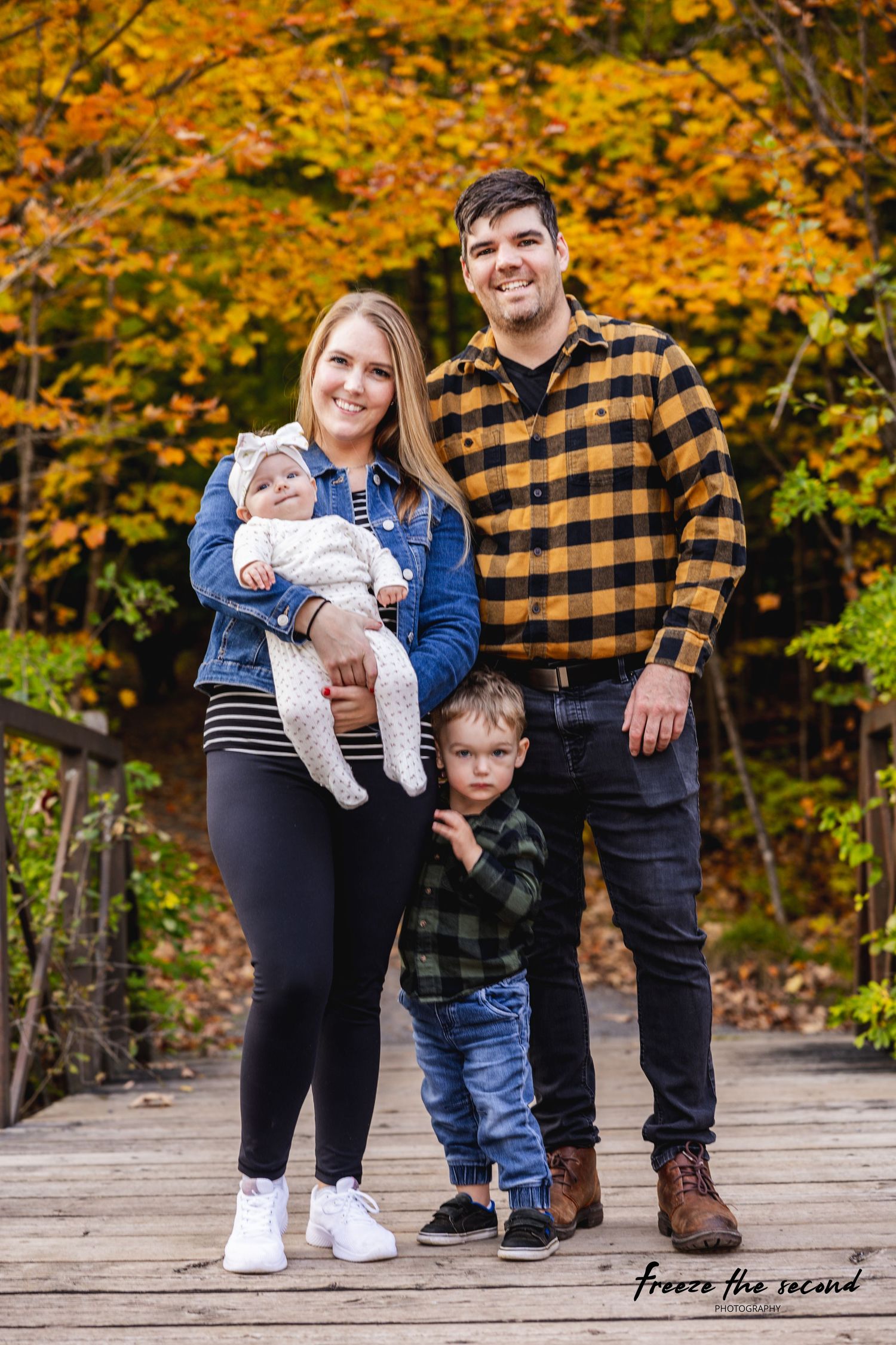Smiling family of four poses on wooden bridge surrounded by vibrant fall foliage.
