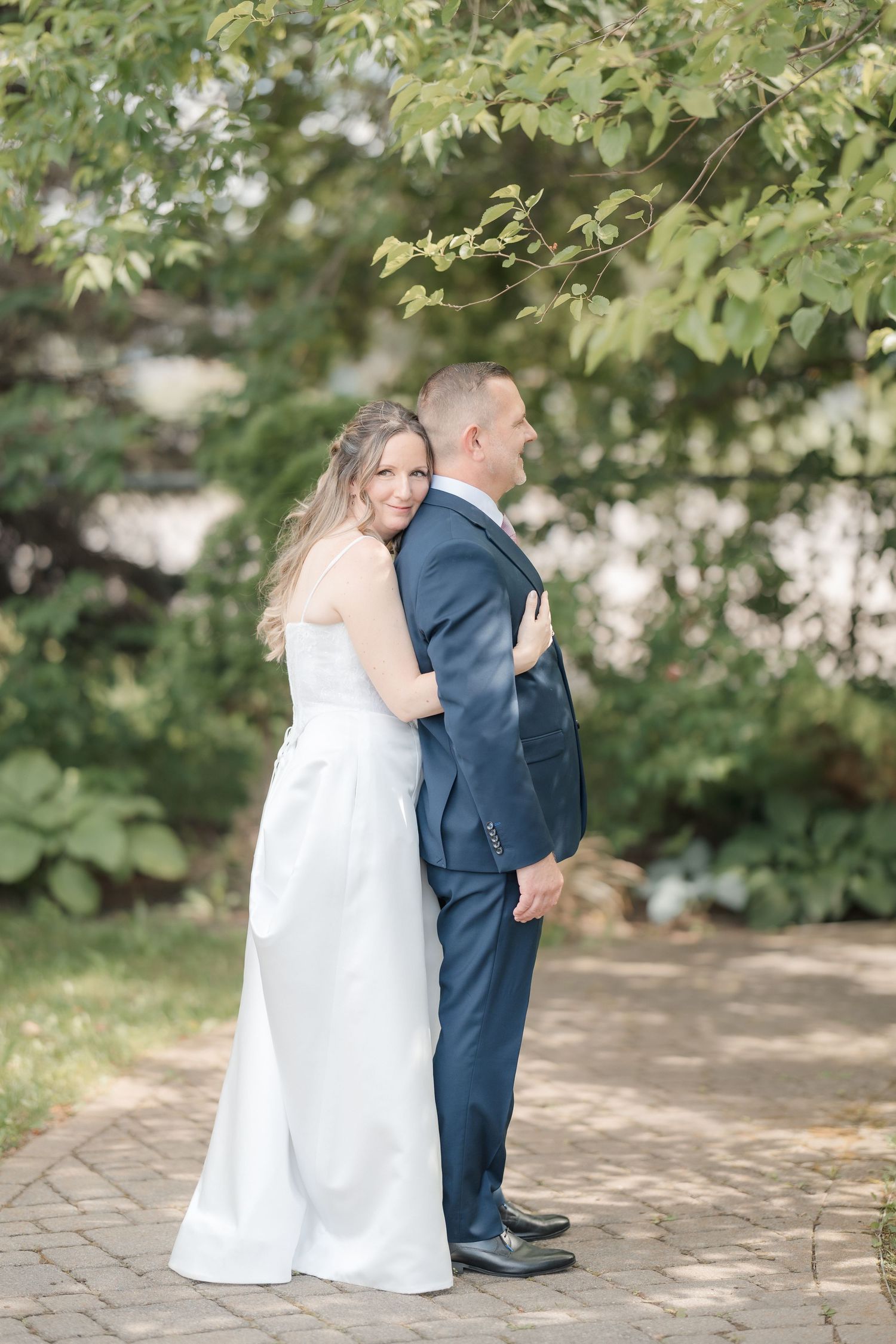 Bride in white gown embraces groom in navy suit from behind on a brick path surrounded by lush greenery.