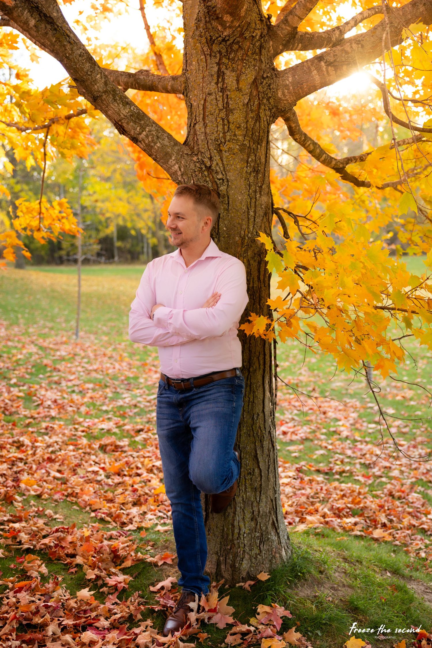 Man in pink shirt leans against large maple tree surrounded by vibrant autumn foliage.