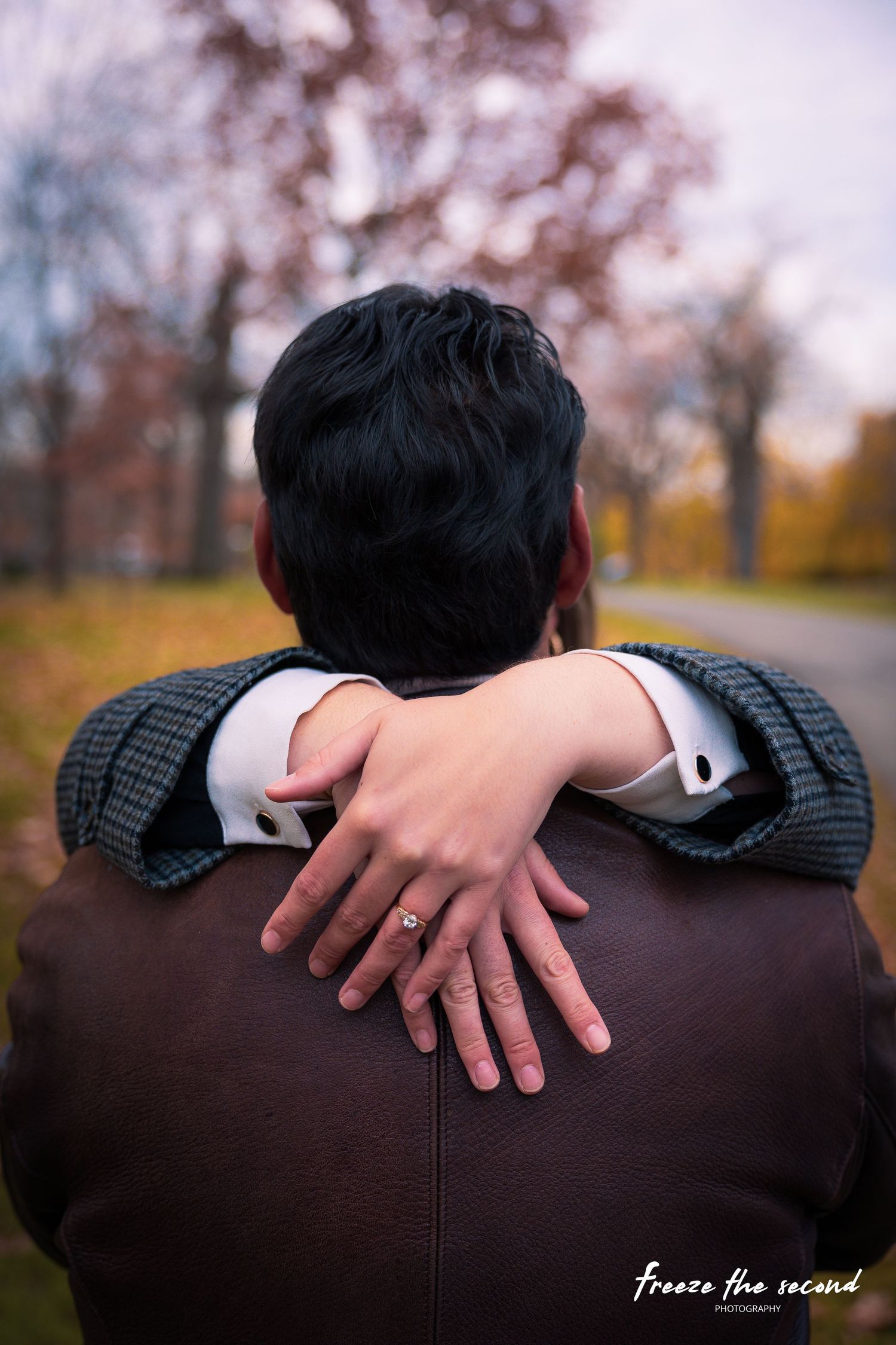 Couple embracing from behind in autumn park, woman's hand showing engagement ring on man's shoulder.