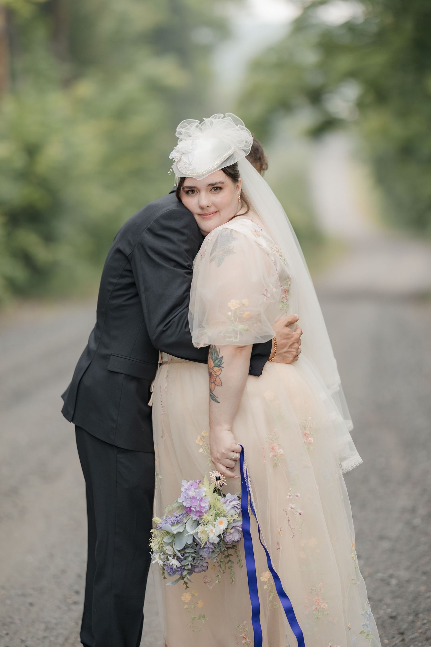 Bride in blush gown with veil and bouquet embraces groom in black suit on a tree-lined road.