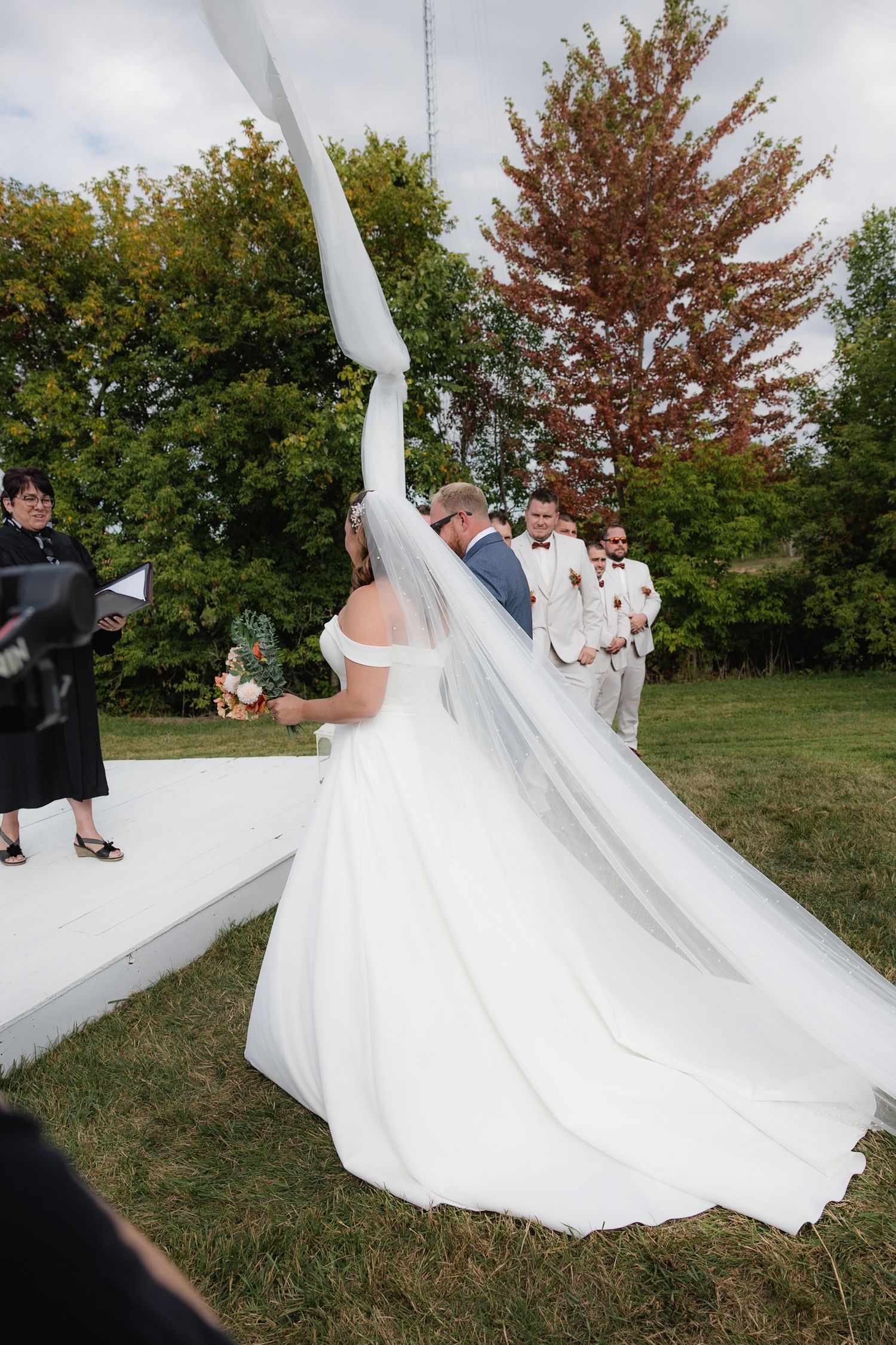 Bride in white off-shoulder gown with flowing veil at outdoor wedding ceremony with groomsmen waiting at altar.
