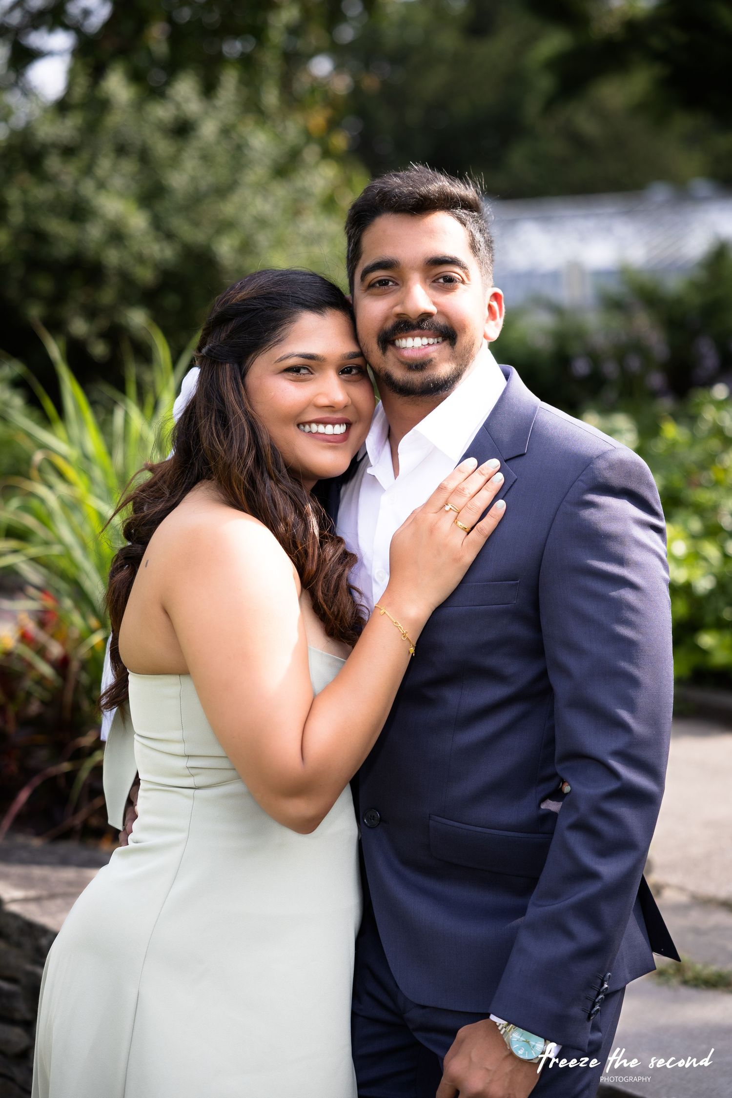 Smiling couple posing together in lush garden, woman in white dress leaning on man in navy suit.