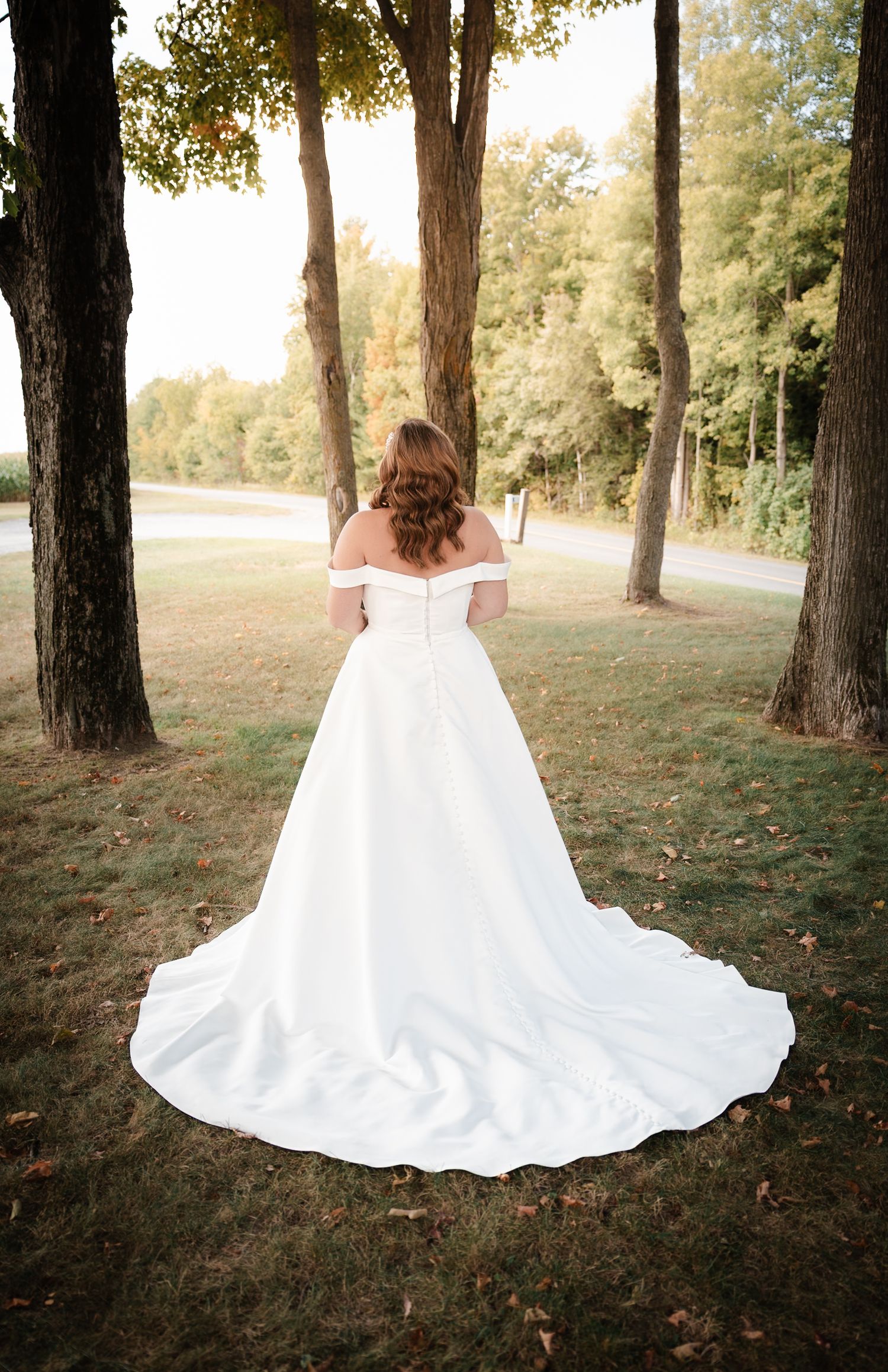 Bride in off-shoulder white ball gown with long train stands between trees in lush green outdoor setting.