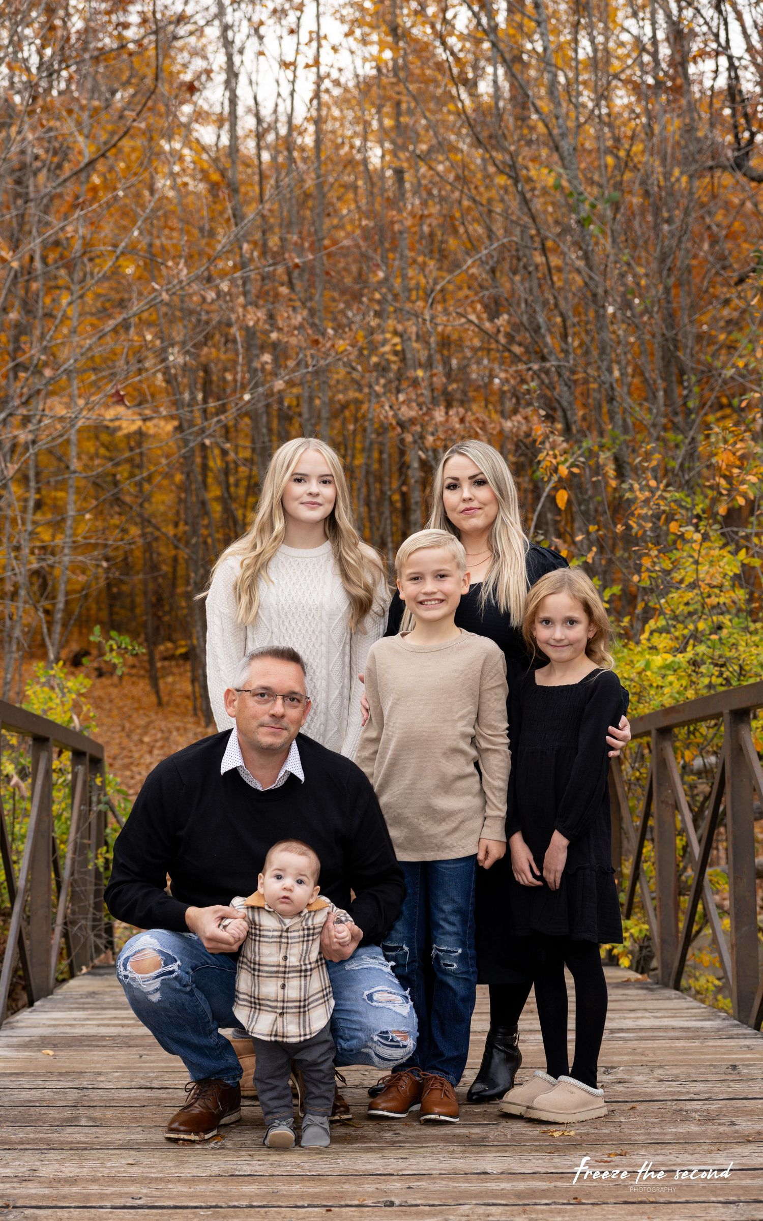 A family of five poses on a wooden bridge surrounded by colorful autumn trees.