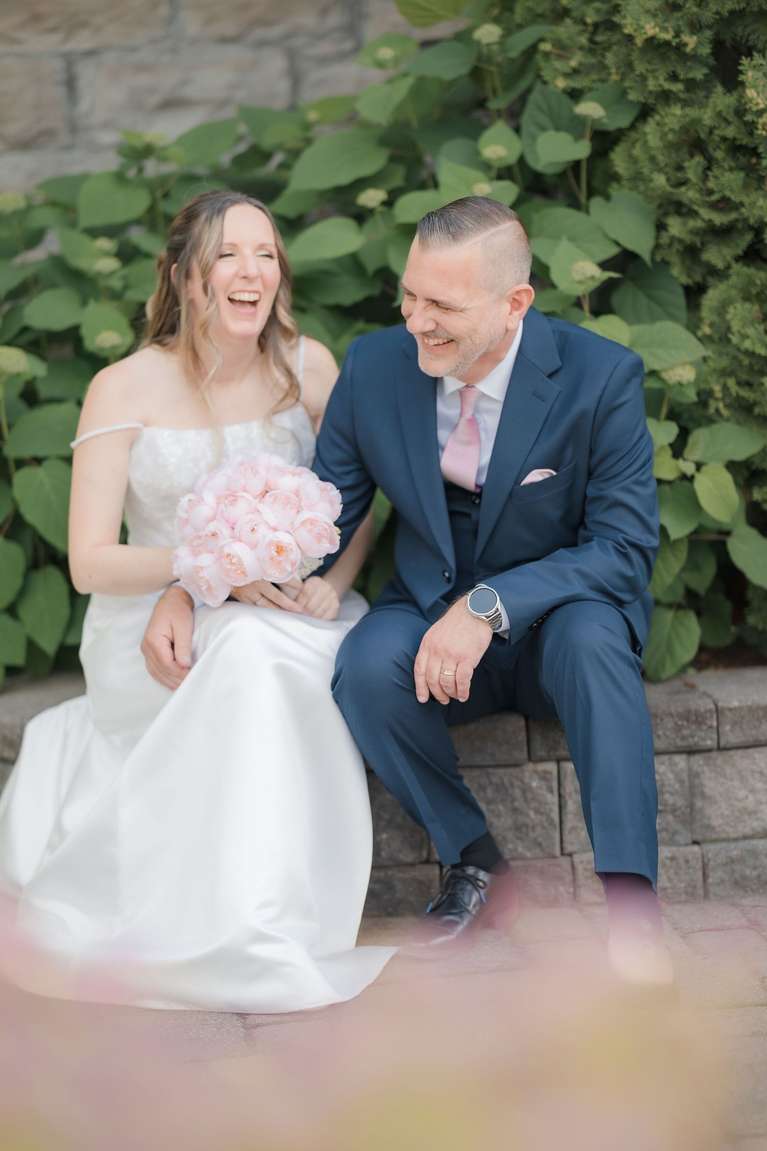 Laughing bride holding pink bouquet and groom in navy suit sitting together on stone steps outdoors.