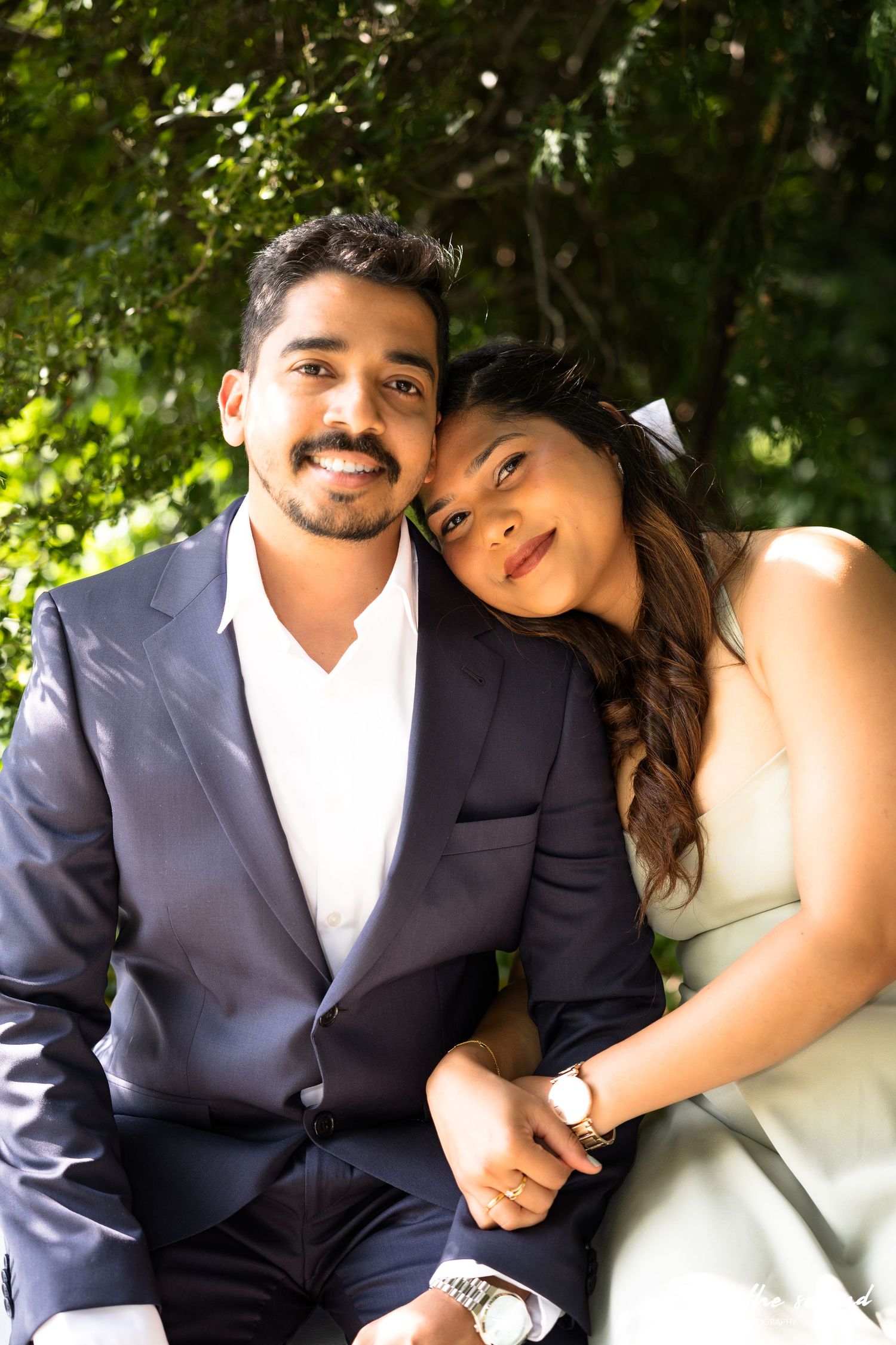 Couple sitting together under green trees, woman resting head on man's shoulder in sunlight.