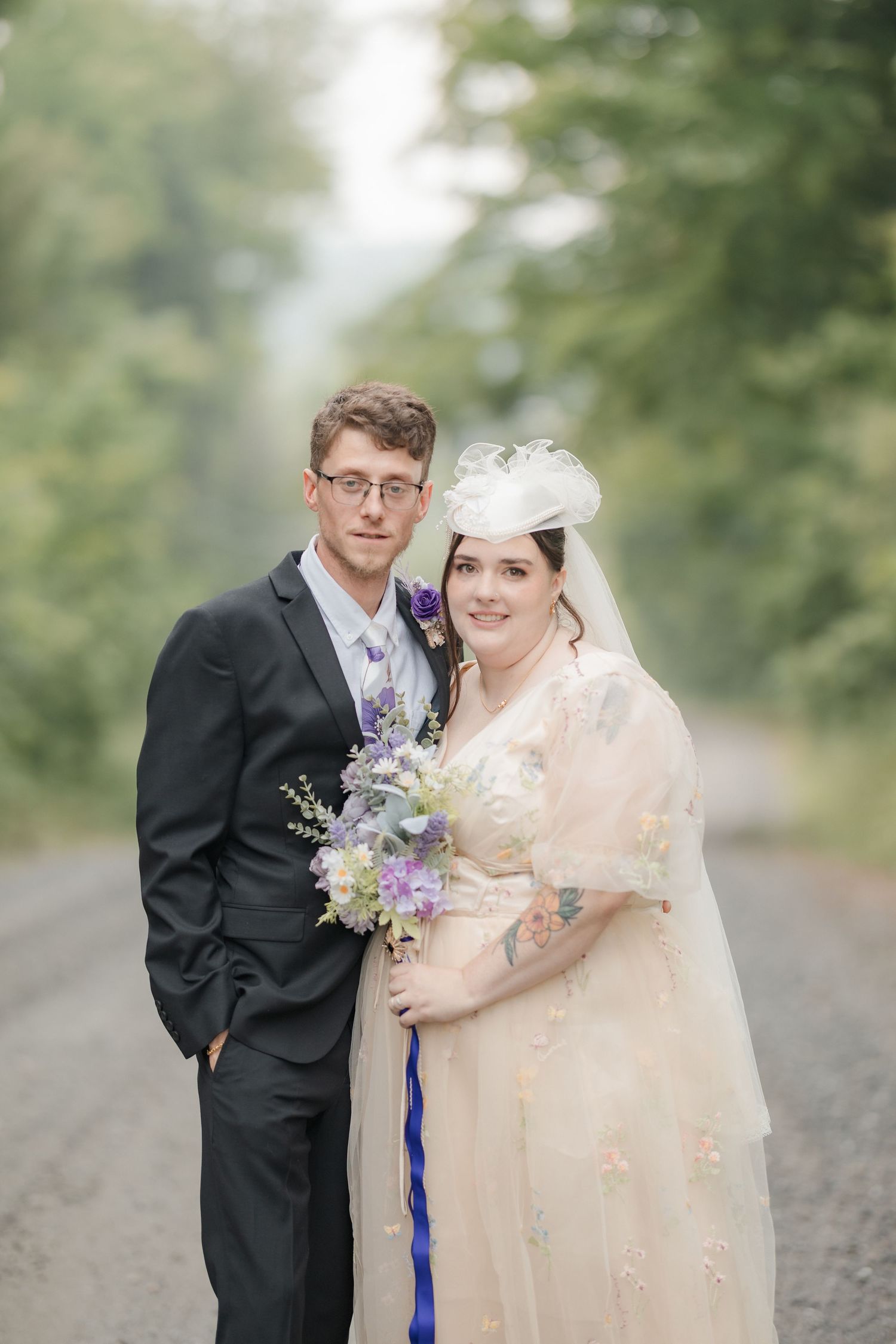 A bride and groom pose on a misty tree-lined road, she holds purple flowers wearing a vintage lace gown with veil.