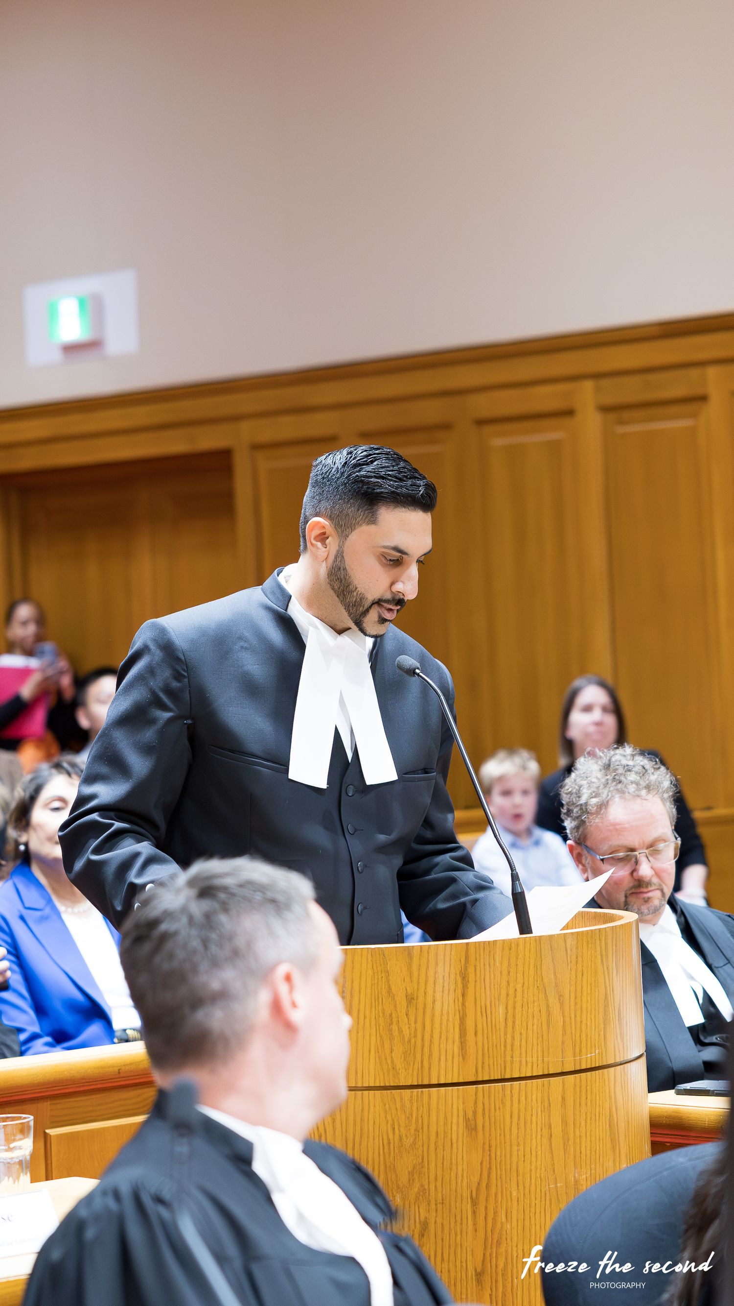 Lawyer in traditional black robes and white tabs speaking at courtroom podium during ceremony.