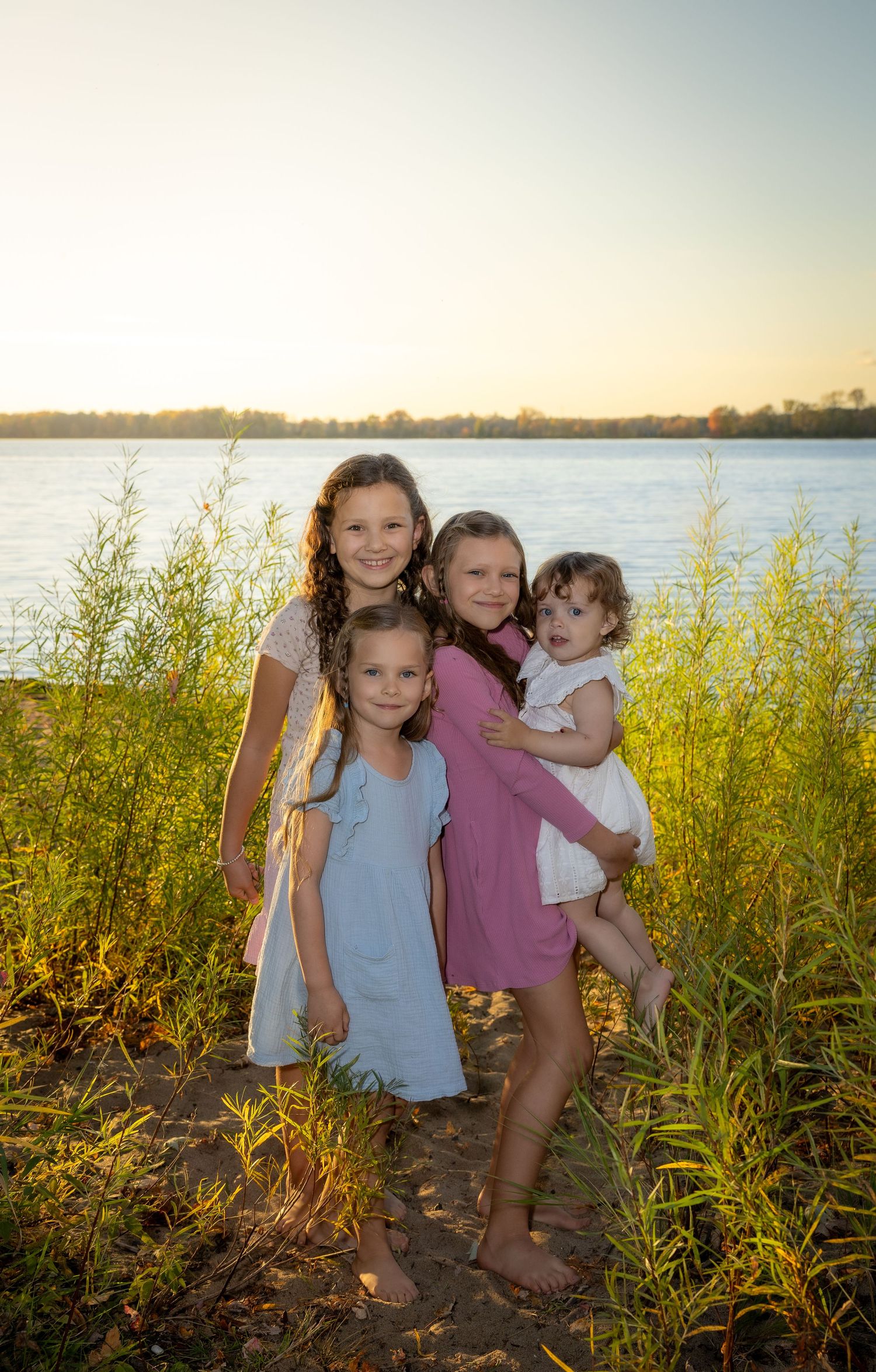 Four sisters pose together in dresses near a sunlit lake at golden hour.