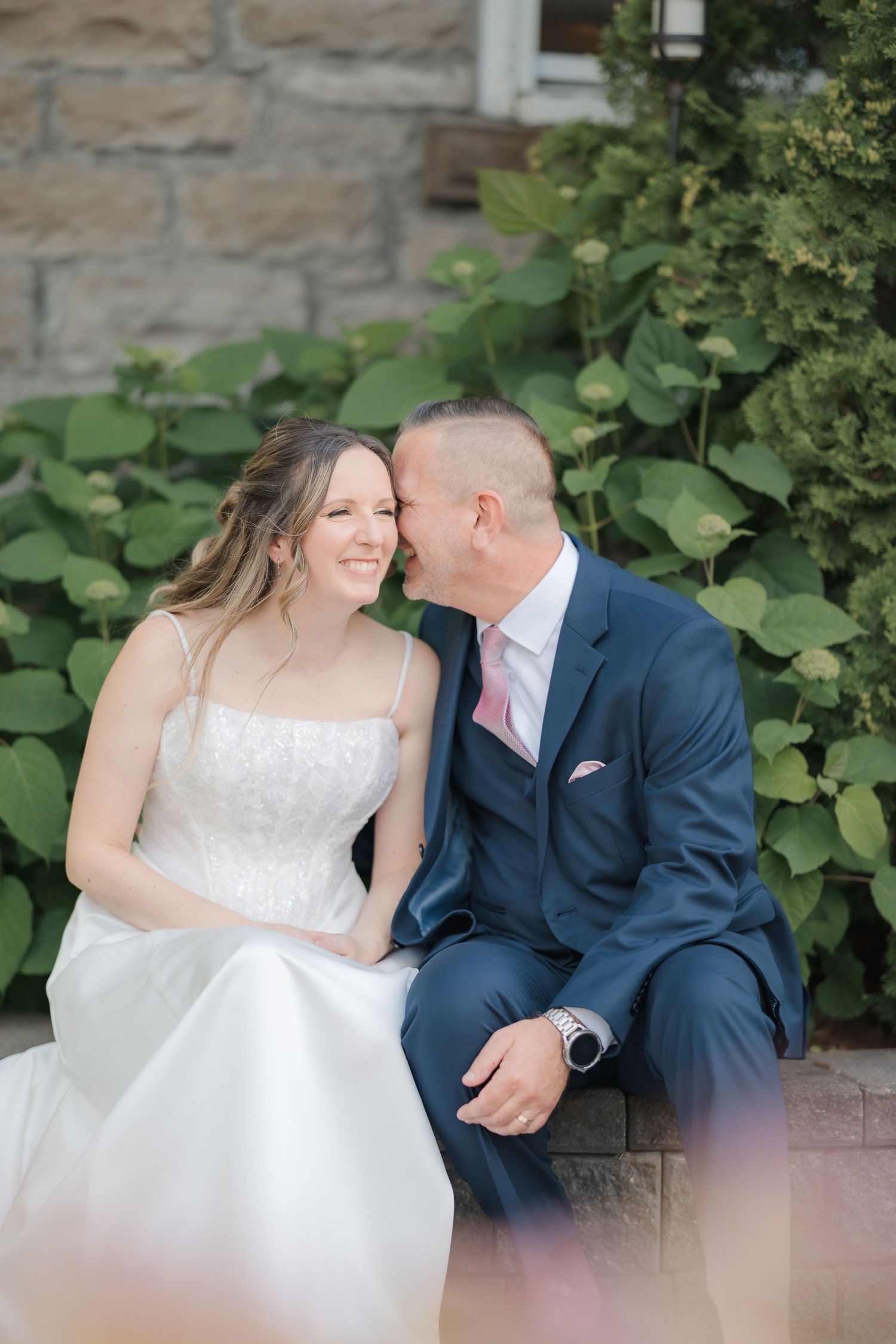 Groom in navy suit whispering to smiling bride in white dress seated on stone steps with greenery.