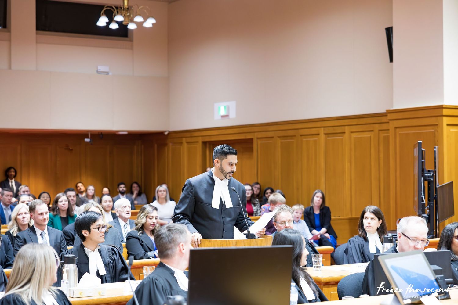 Lawyer in black robes addresses a full courtroom audience during a formal legal proceeding.