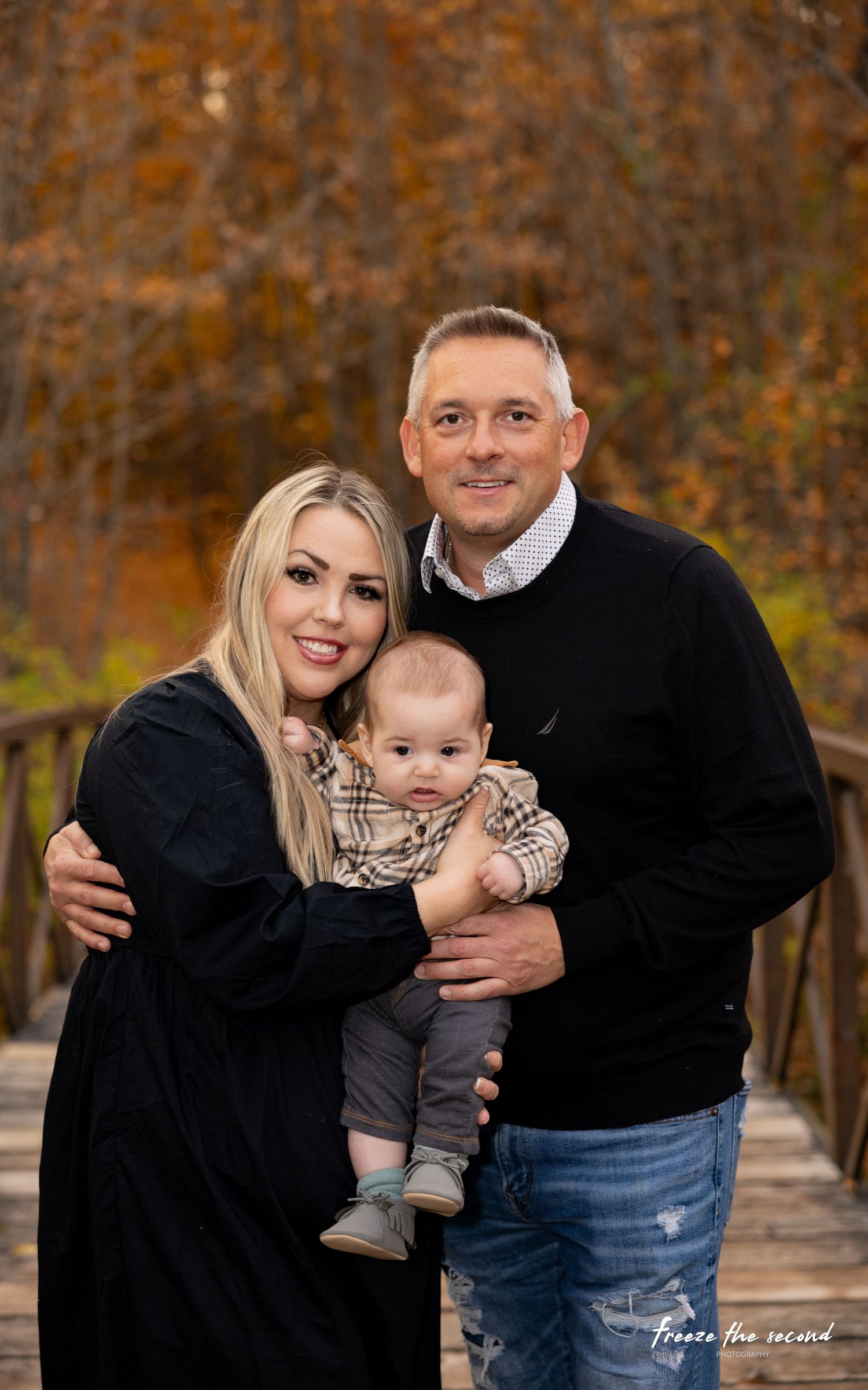 A smiling couple holds their baby on a wooden bridge with fall foliage behind them.