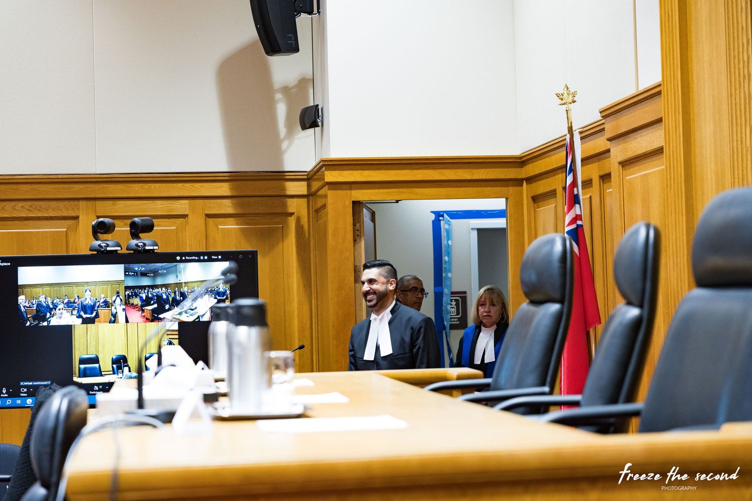 Smiling lawyer in robes stands in courtroom with attendees visible on video screen in background.