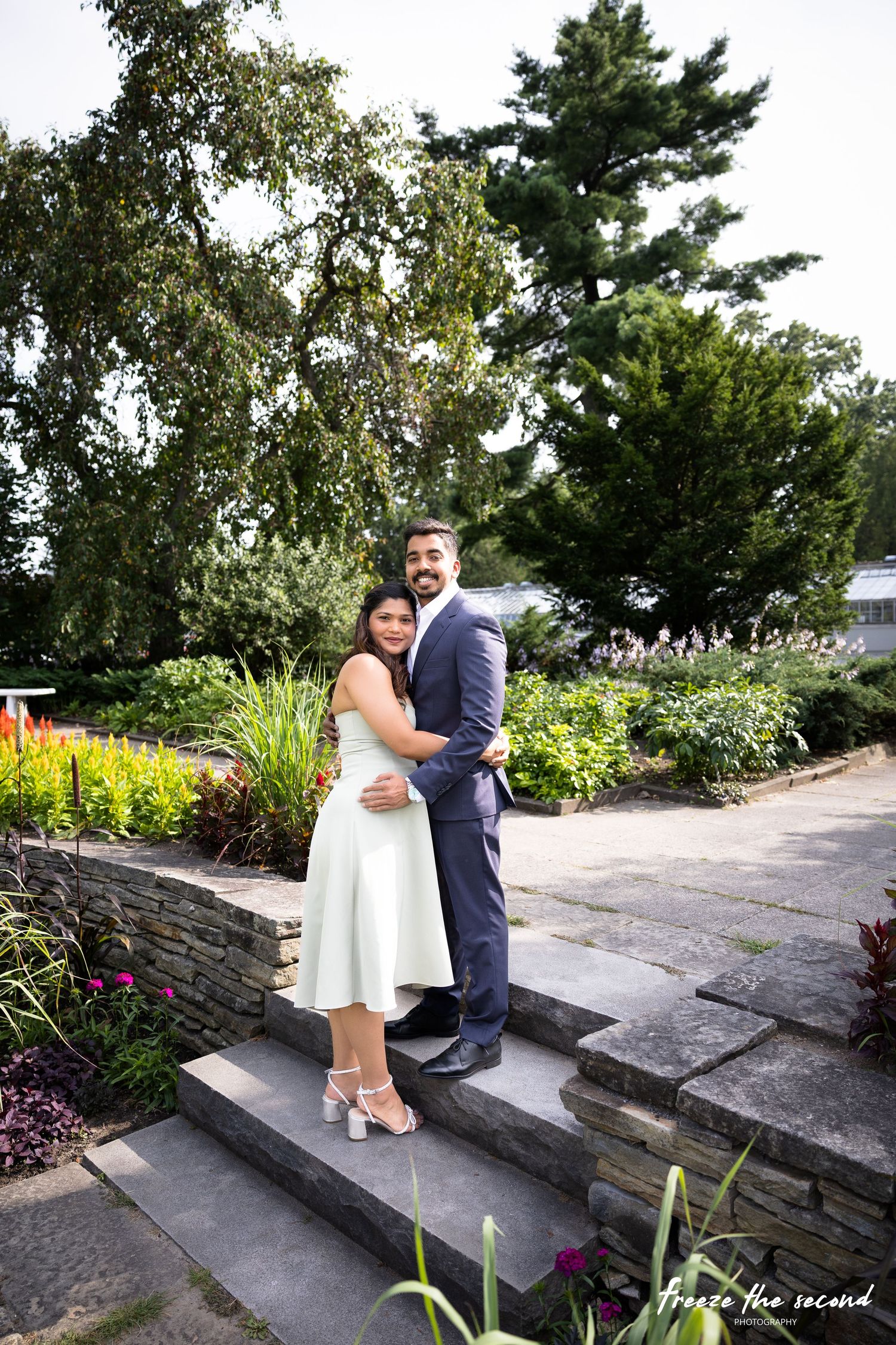 Elegant couple embracing on garden steps surrounded by colorful flowers and green trees.
