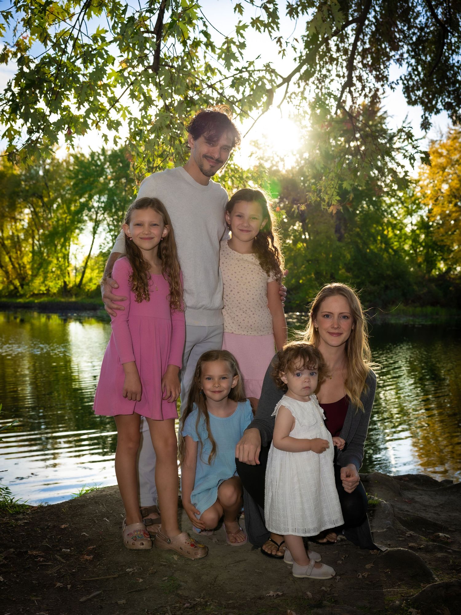 A family of six smiles together near a sunlit pond surrounded by green trees.