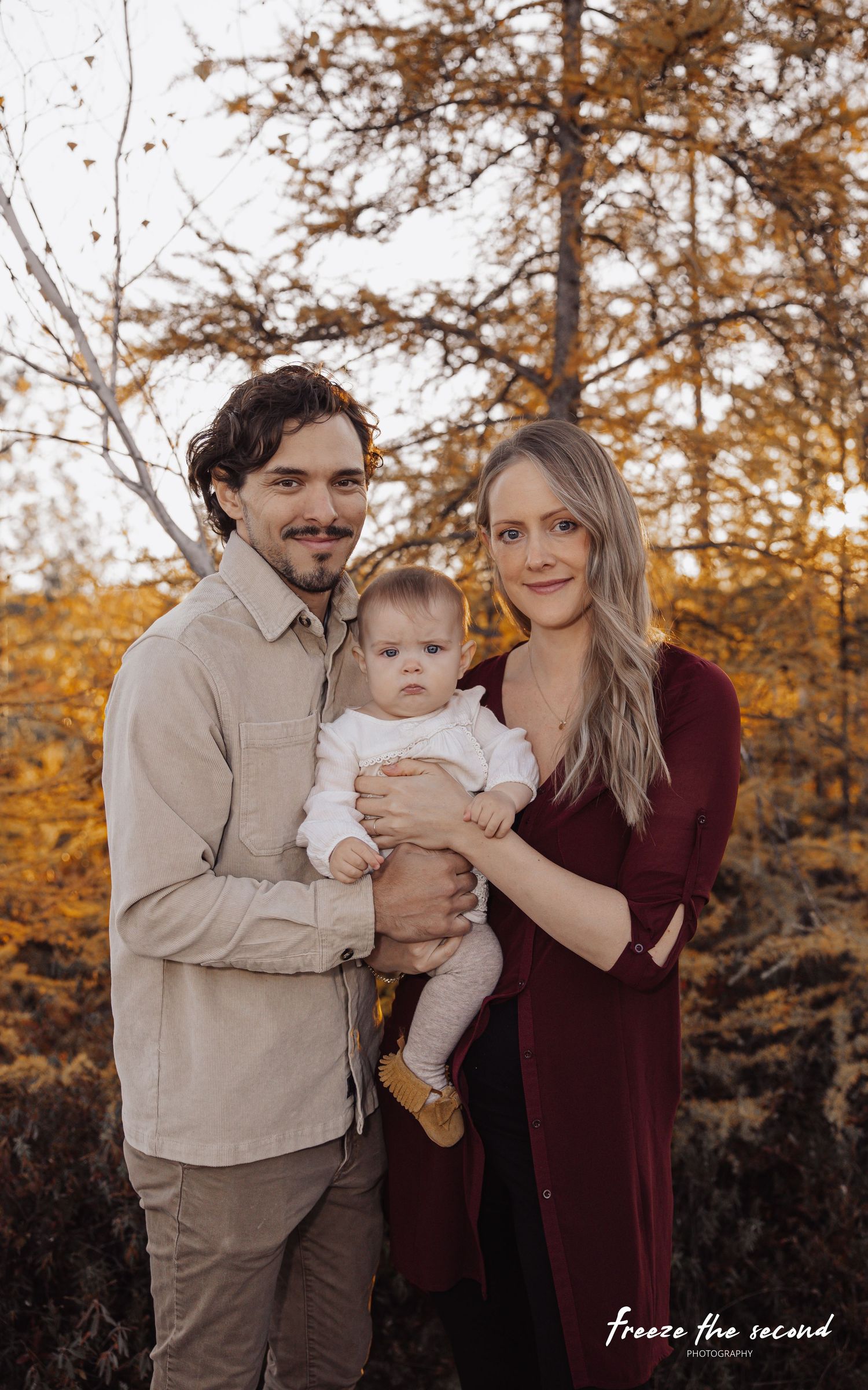 Young couple holds baby during warm autumn family portrait among golden larch trees.