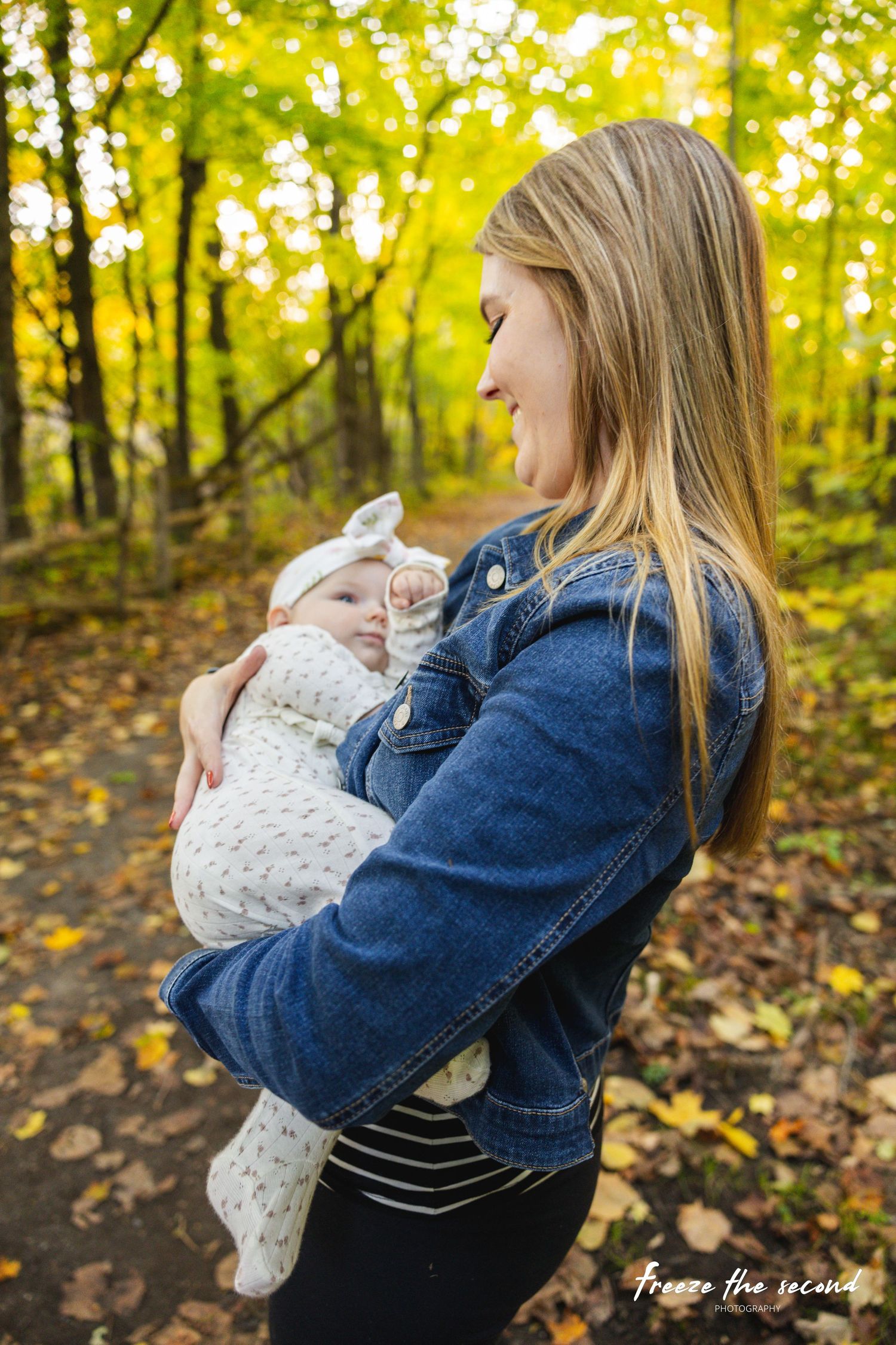 Smiling mother in denim jacket cradles newborn baby in autumn forest park setting.