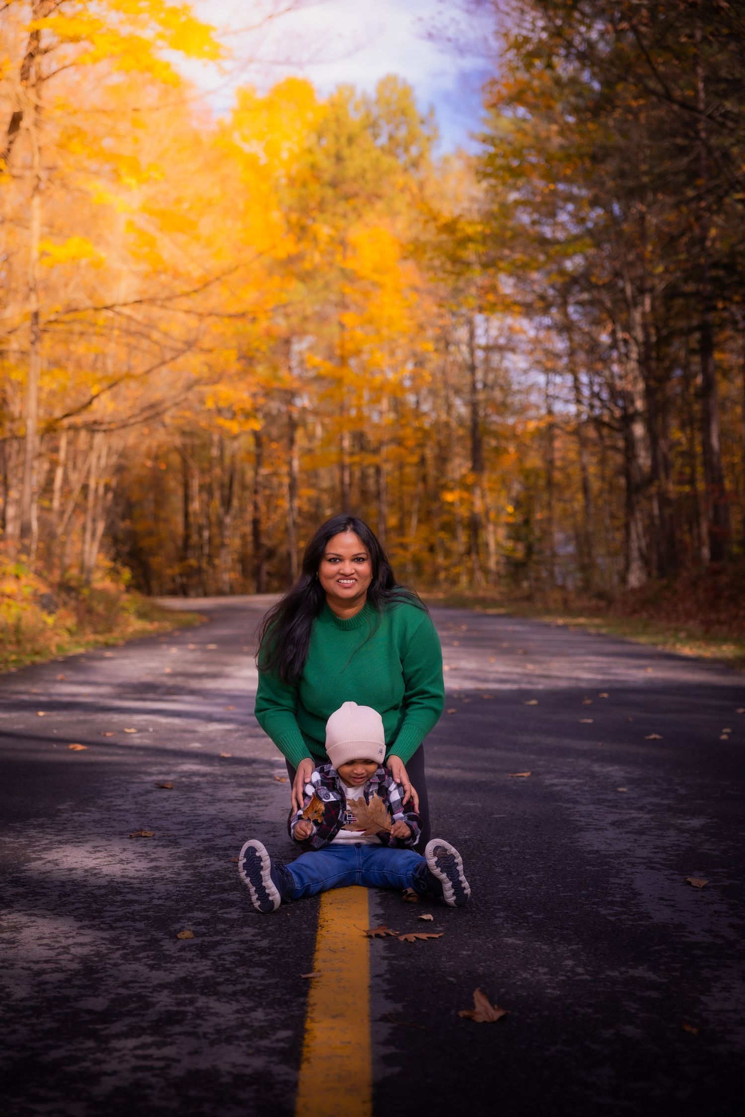 Mother in green sweater sits on road centerline with toddler amid vibrant fall trees.