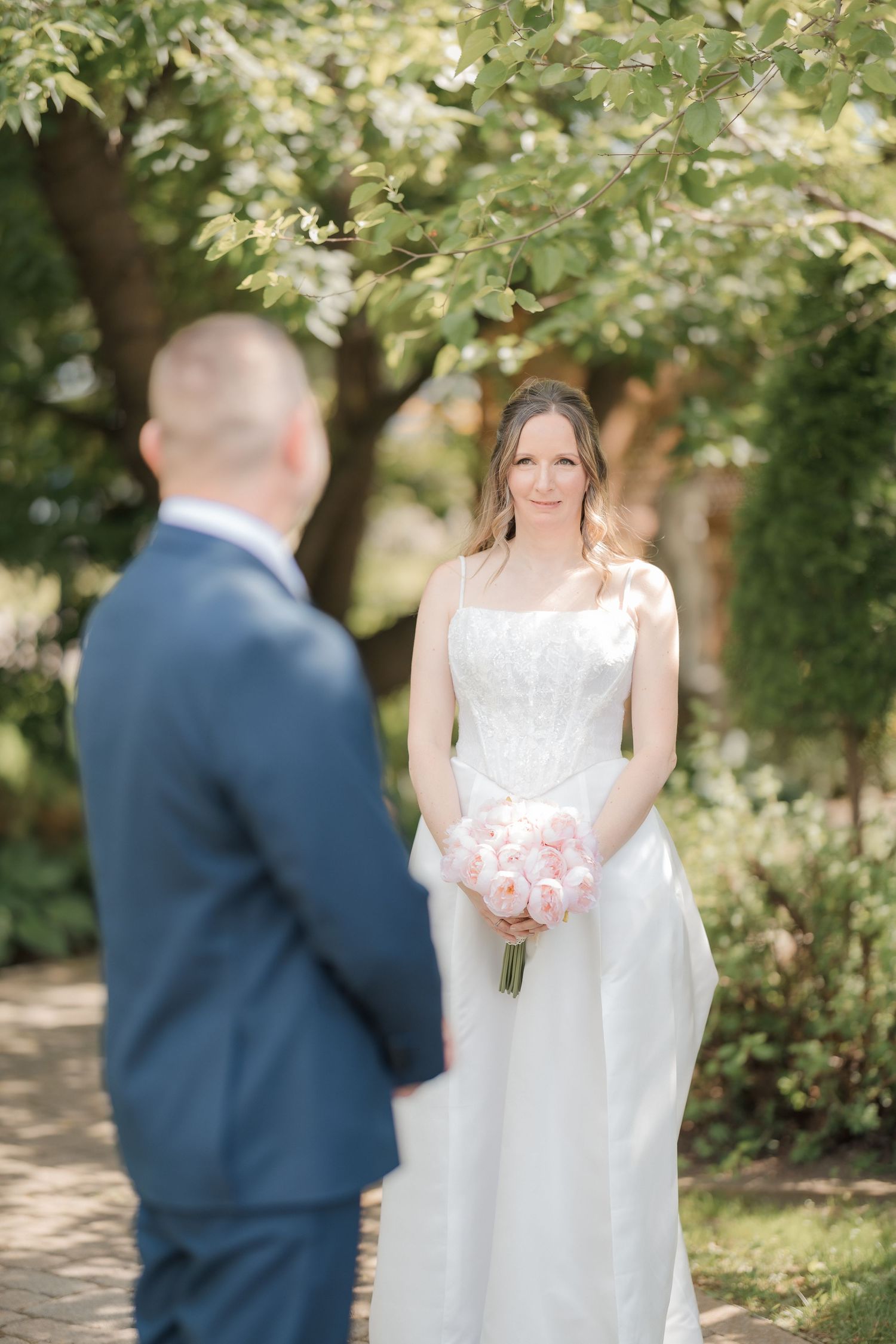 Bride in white gown holding pink bouquet smiling at groom during first look.