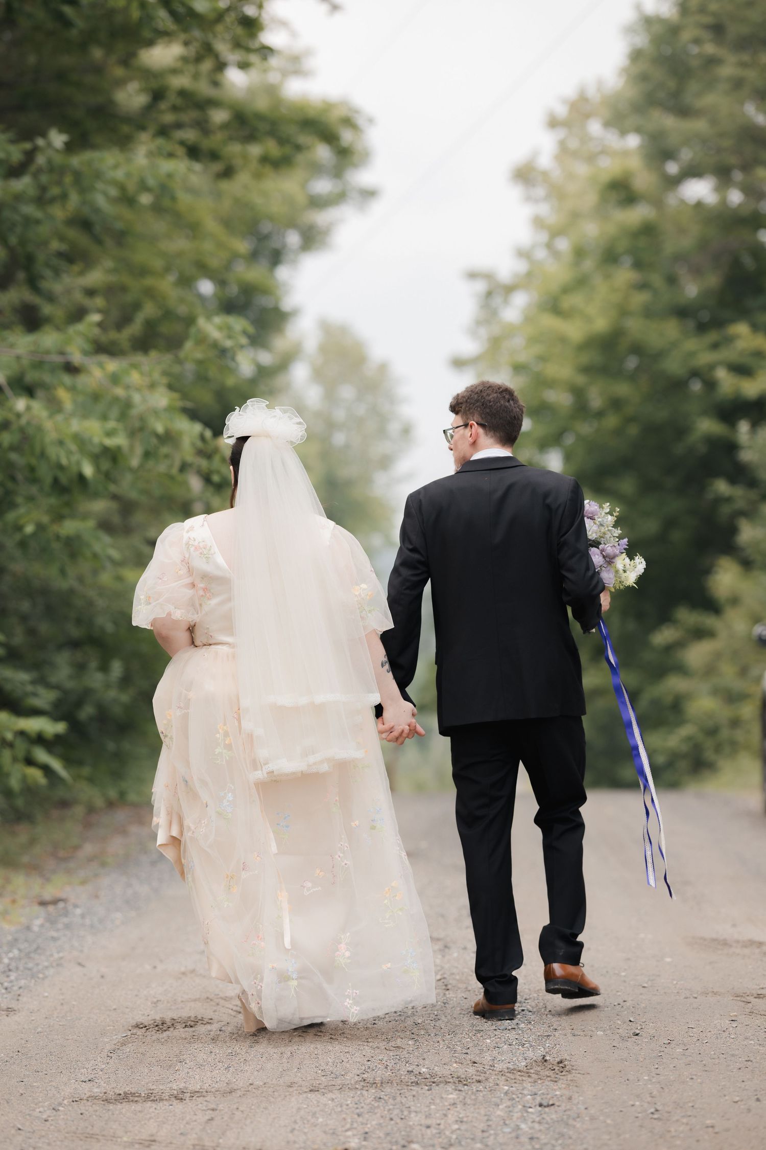 Bride and groom walking hand in hand down a tree-lined path, viewed from behind on their wedding day.