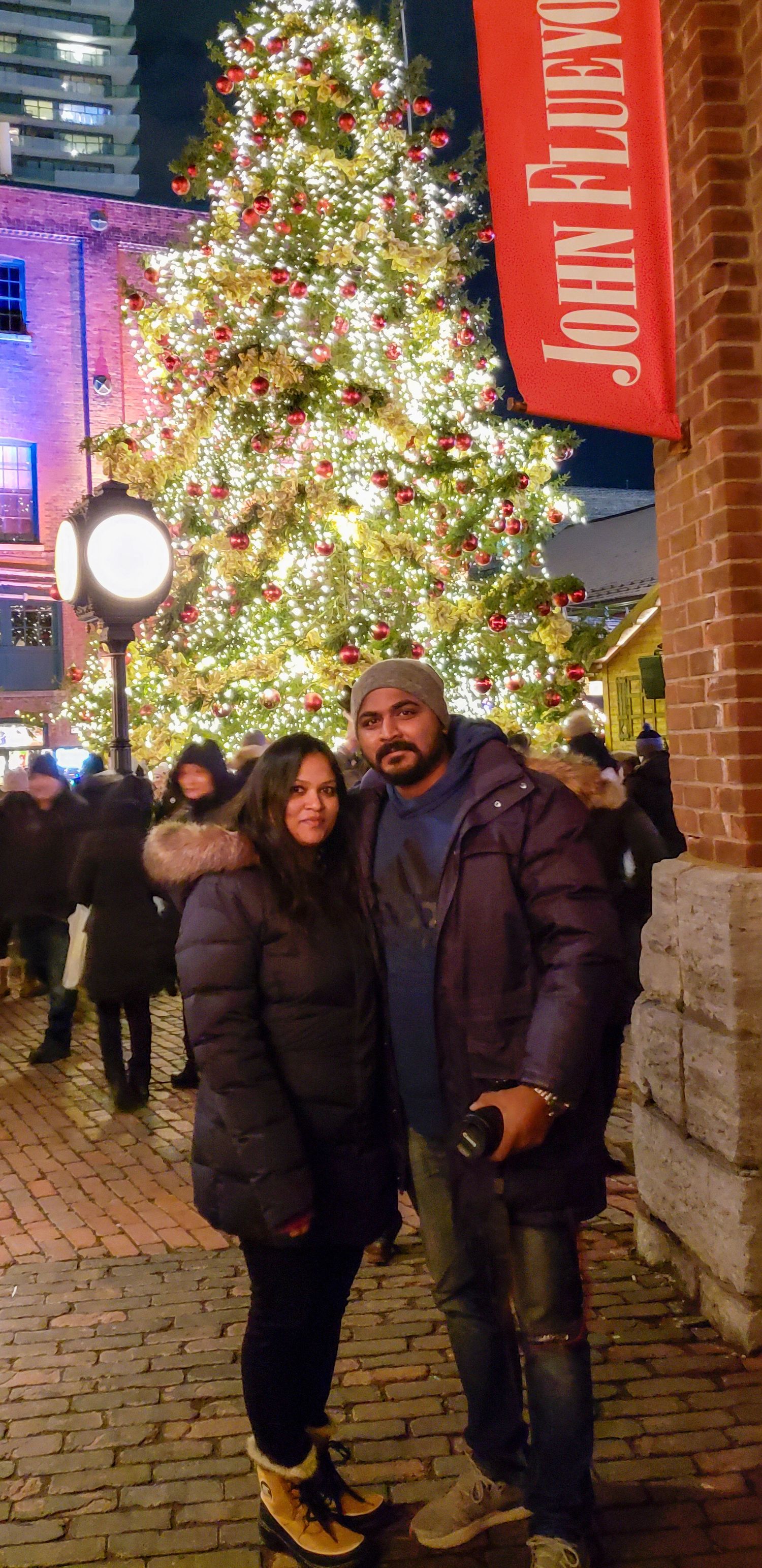 Couple posing in front of a large decorated Christmas tree at John Fluevog in a festive outdoor setting.