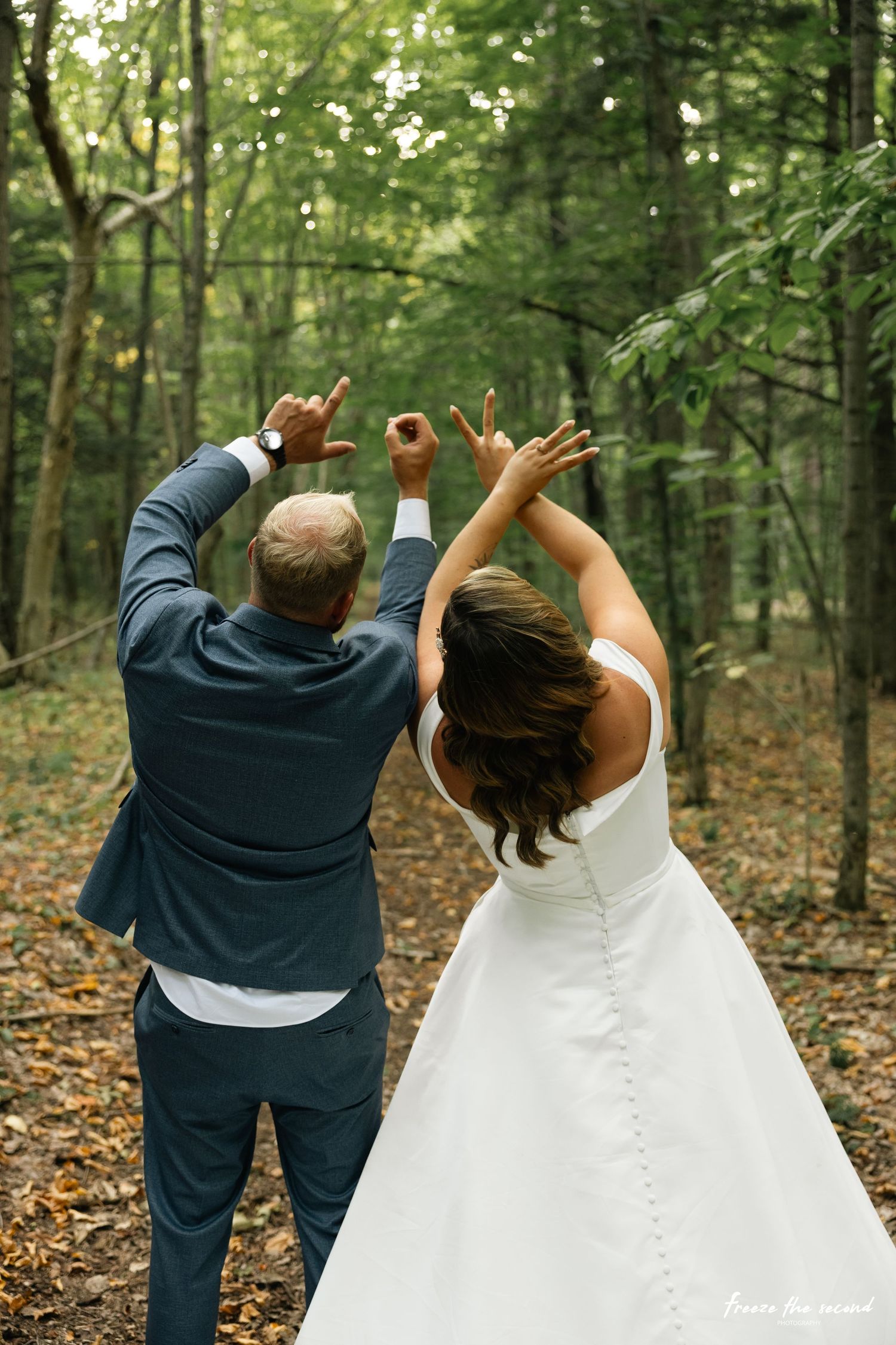 A couple in wedding attire raises their hands together in celebration while standing in a lush green forest.