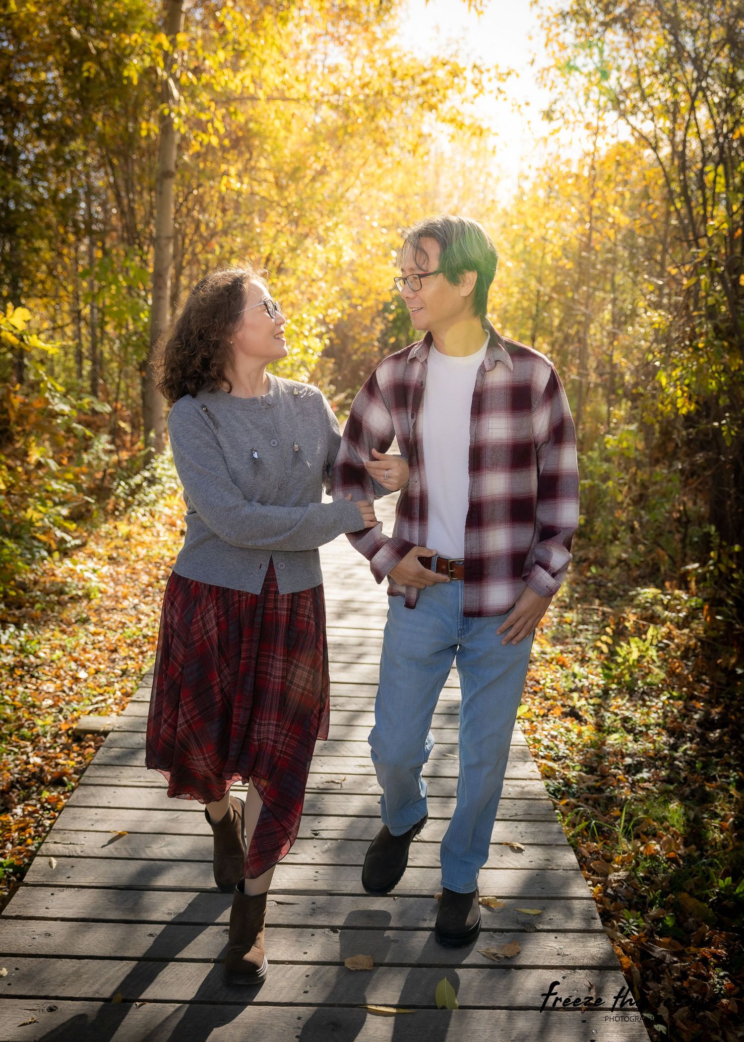 A couple walks hand in hand on a wooden boardwalk through a golden autumn forest.