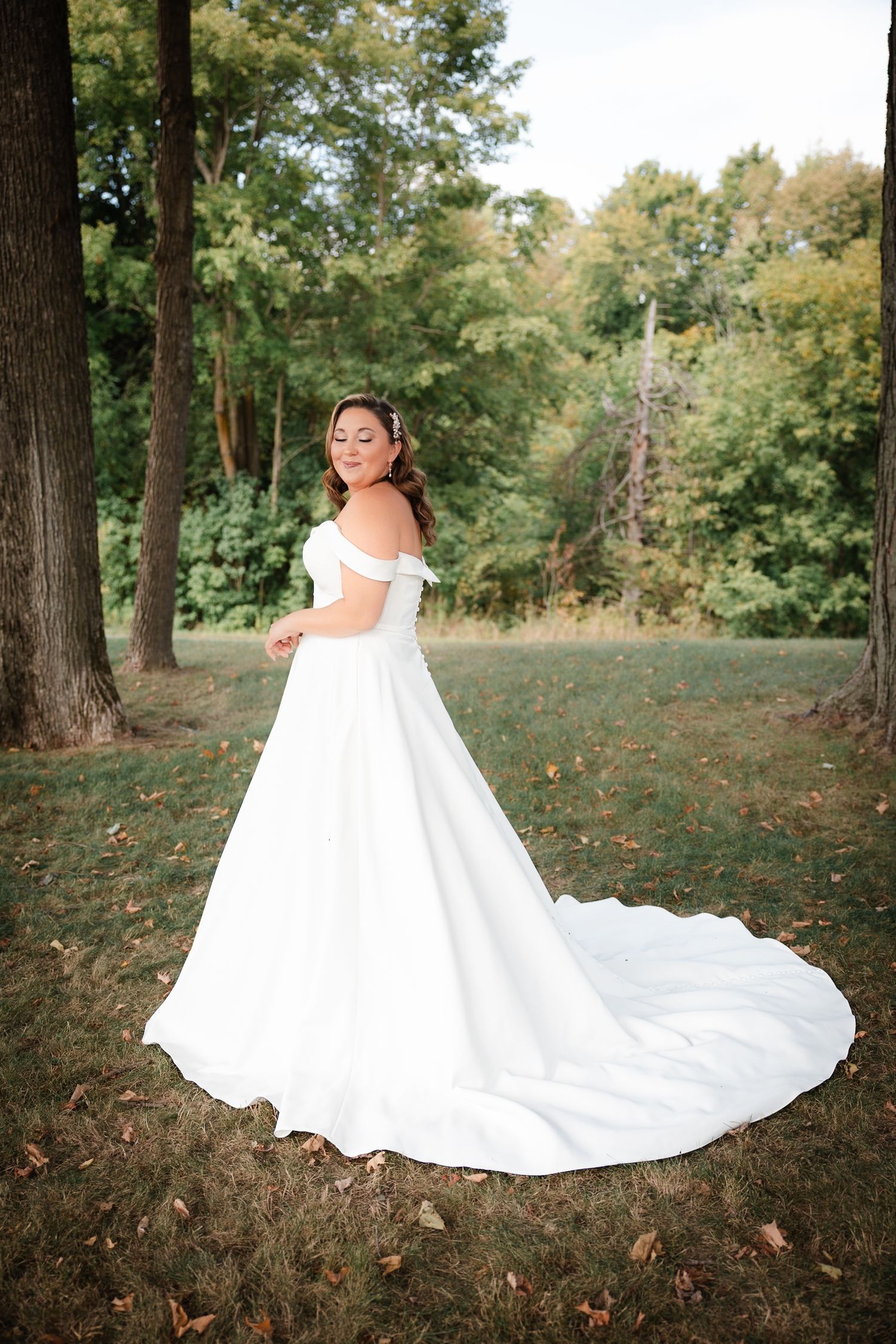 Bride in off-shoulder white satin ball gown with long train posing gracefully in a wooded outdoor setting.
