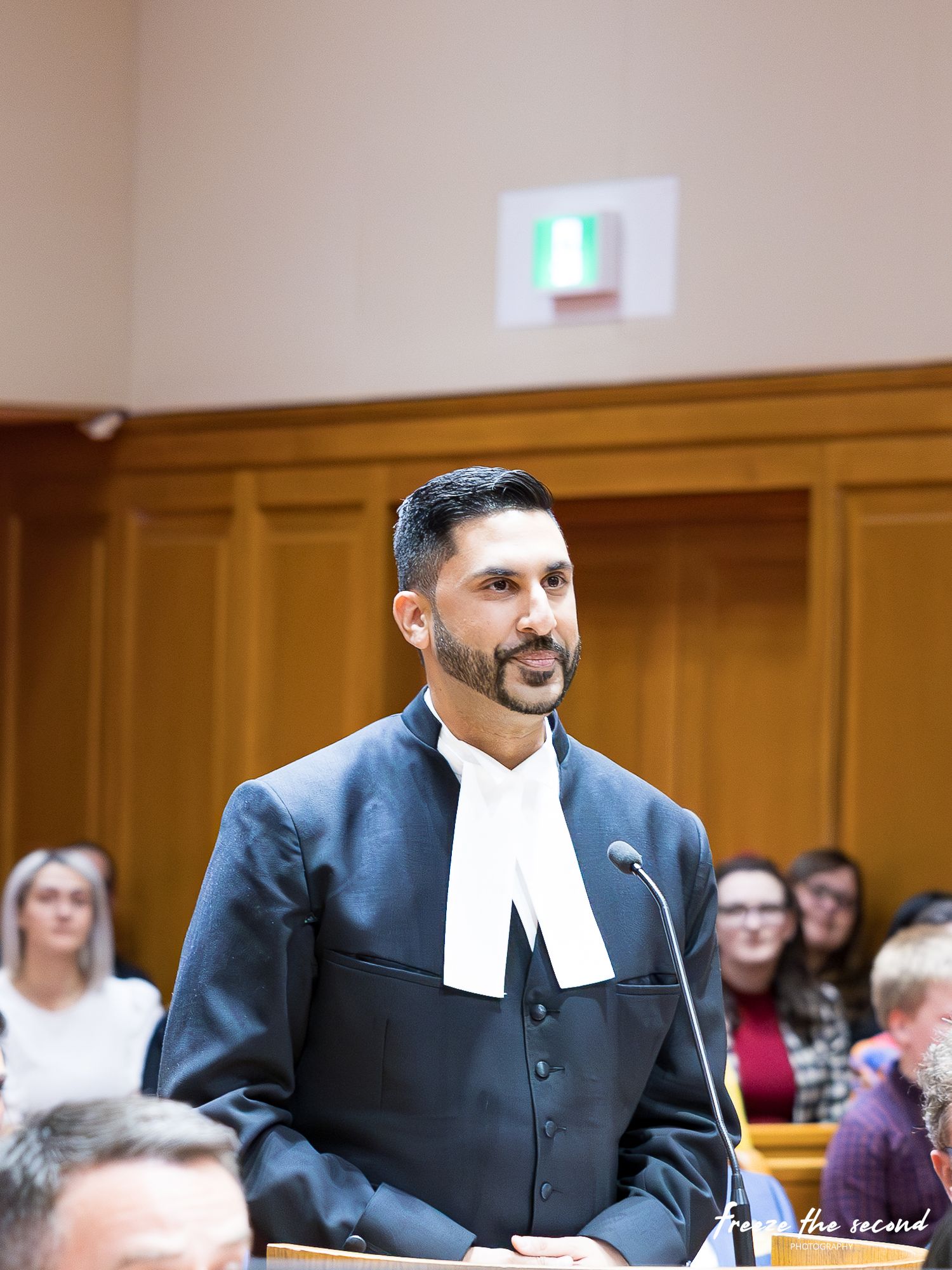 Lawyer in black robes and white jabot stands confidently at podium in formal courtroom during legal ceremony.