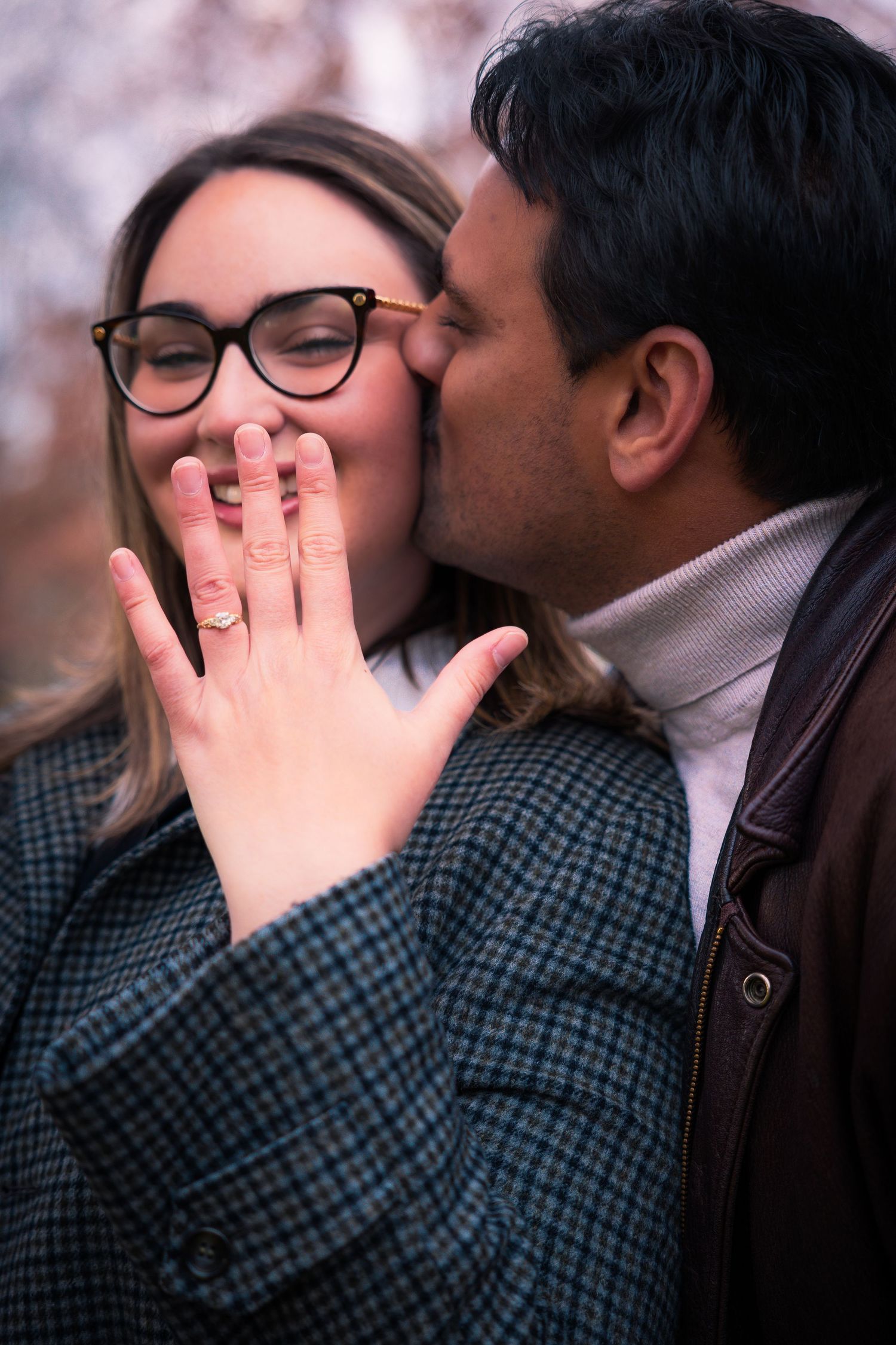 Man kissing woman's cheek as she smiles and displays her engagement ring to the camera.