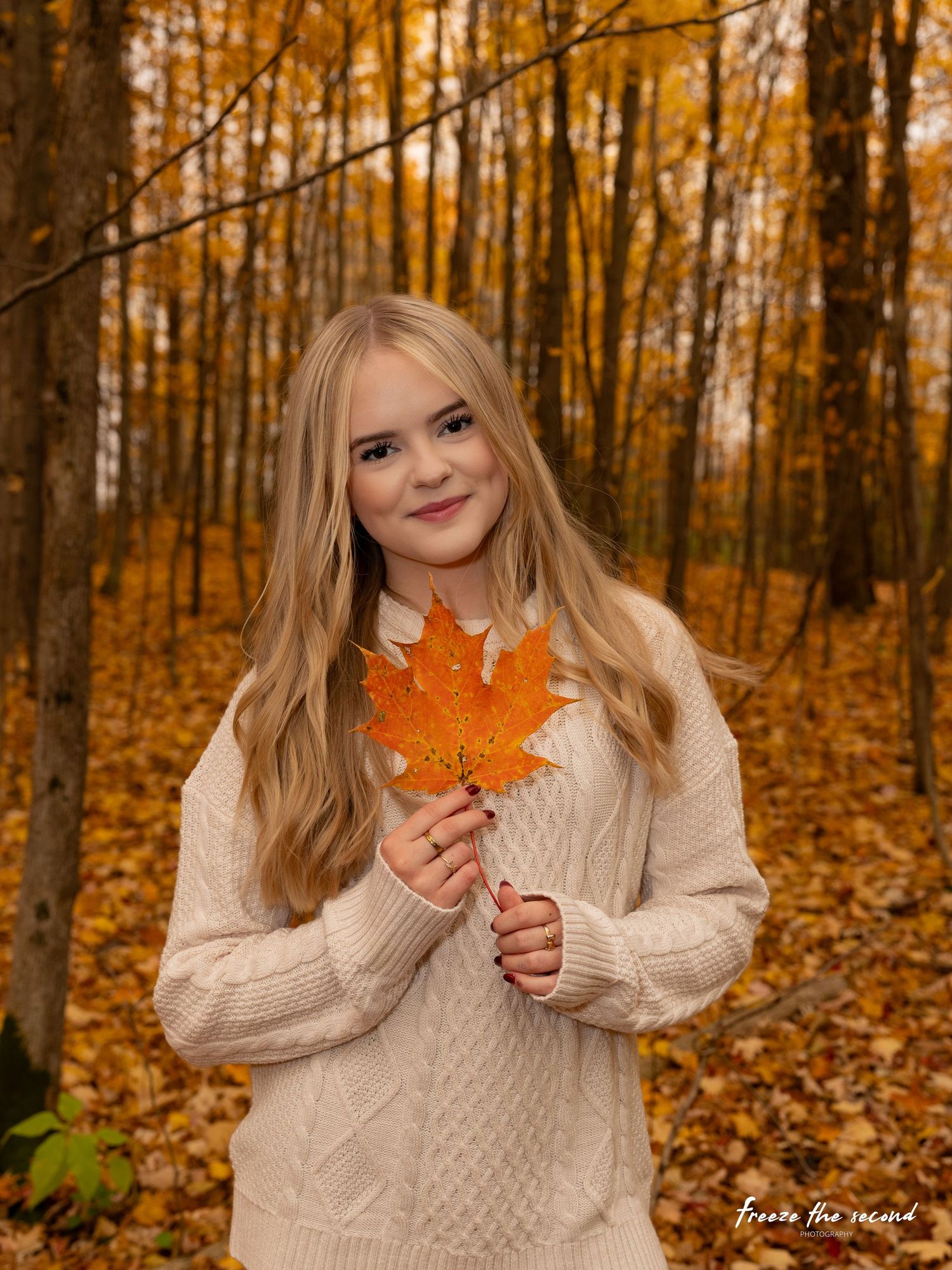 A young blonde girl holds an orange maple leaf in a cozy cream sweater amid fall foliage.