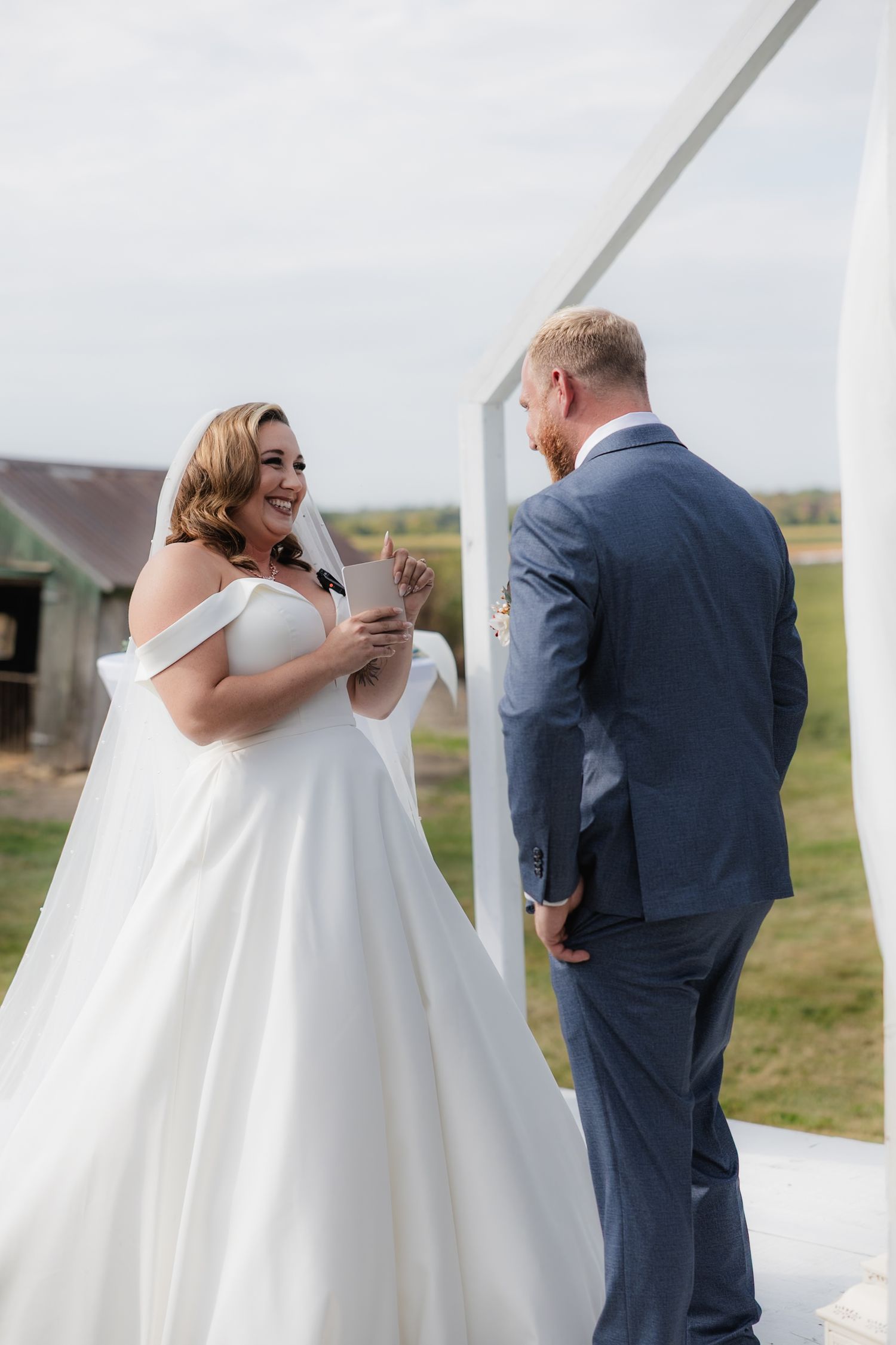 Bride in satin off-shoulder wedding dress reading vows to groom in navy suit at outdoor ceremony.