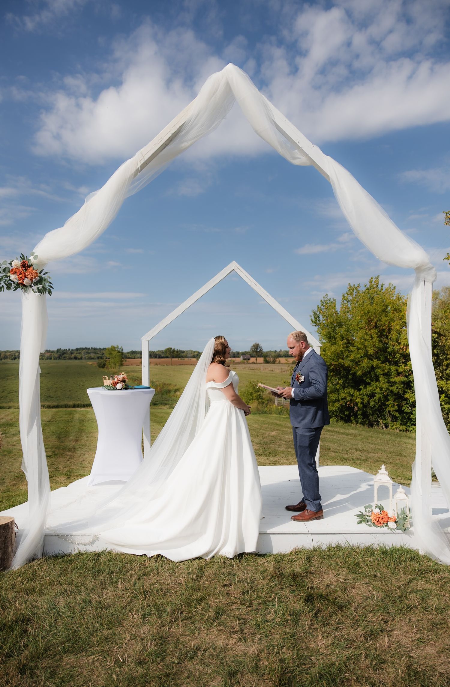 Bride and groom exchanging vows under white geometric arch on sunny outdoor farm ceremony.