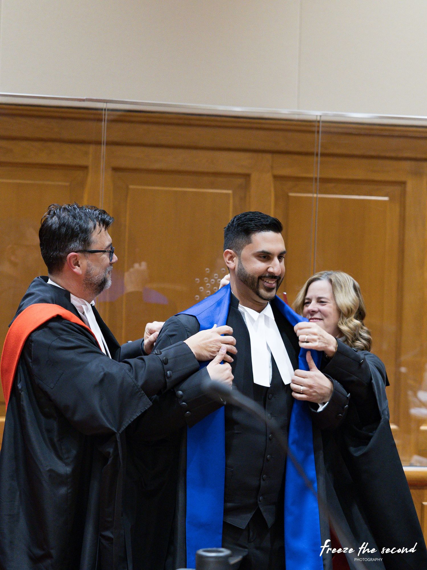 Two robed officials place a blue sash on a smiling lawyer during a bar admission ceremony.
