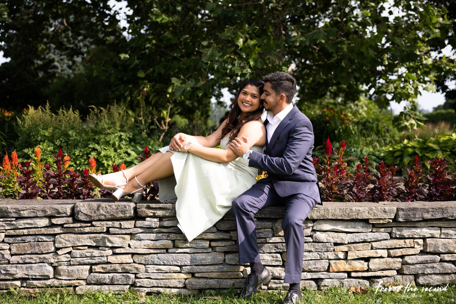 Romantic couple sitting on stone wall in garden, man kissing woman's cheek as she laughs.