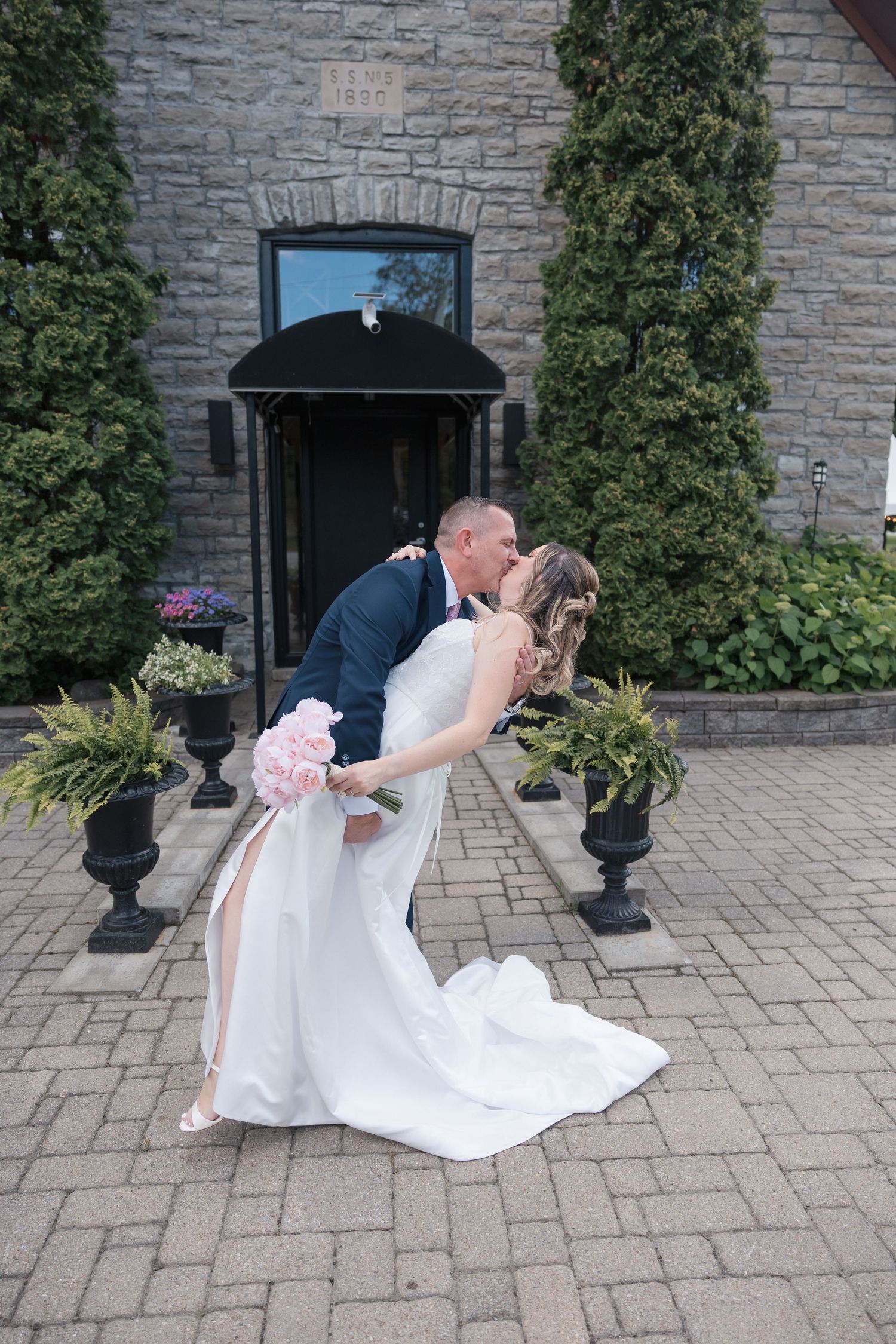 Bride and groom sharing a romantic dip kiss outside a stone building, bride holding pink peony bouquet.