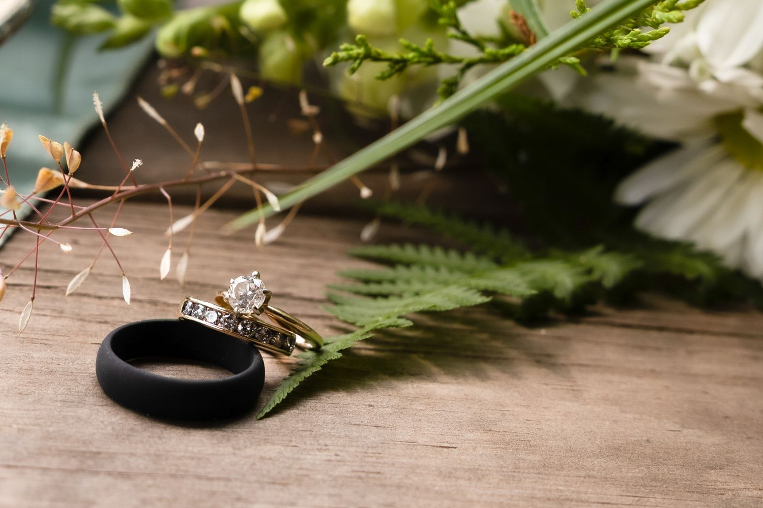 Diamond engagement ring and black silicone wedding band resting on fern leaves beside white flowers at Famoda Farm in Taylors, SC.