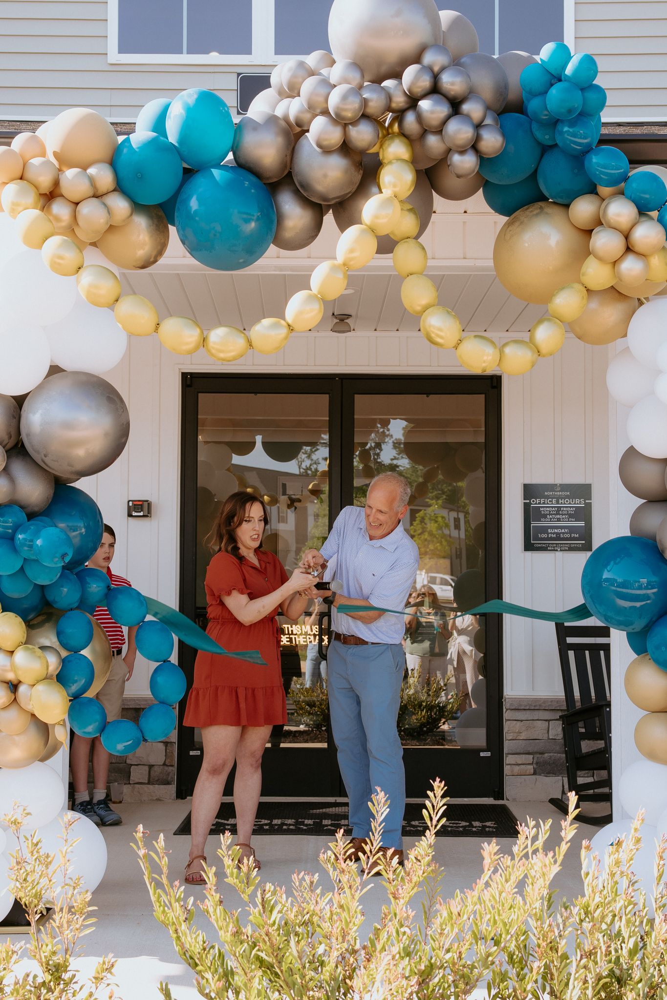 Couple cuts ribbon at grand opening ceremony under colorful teal, gold, and silver balloon arch.
