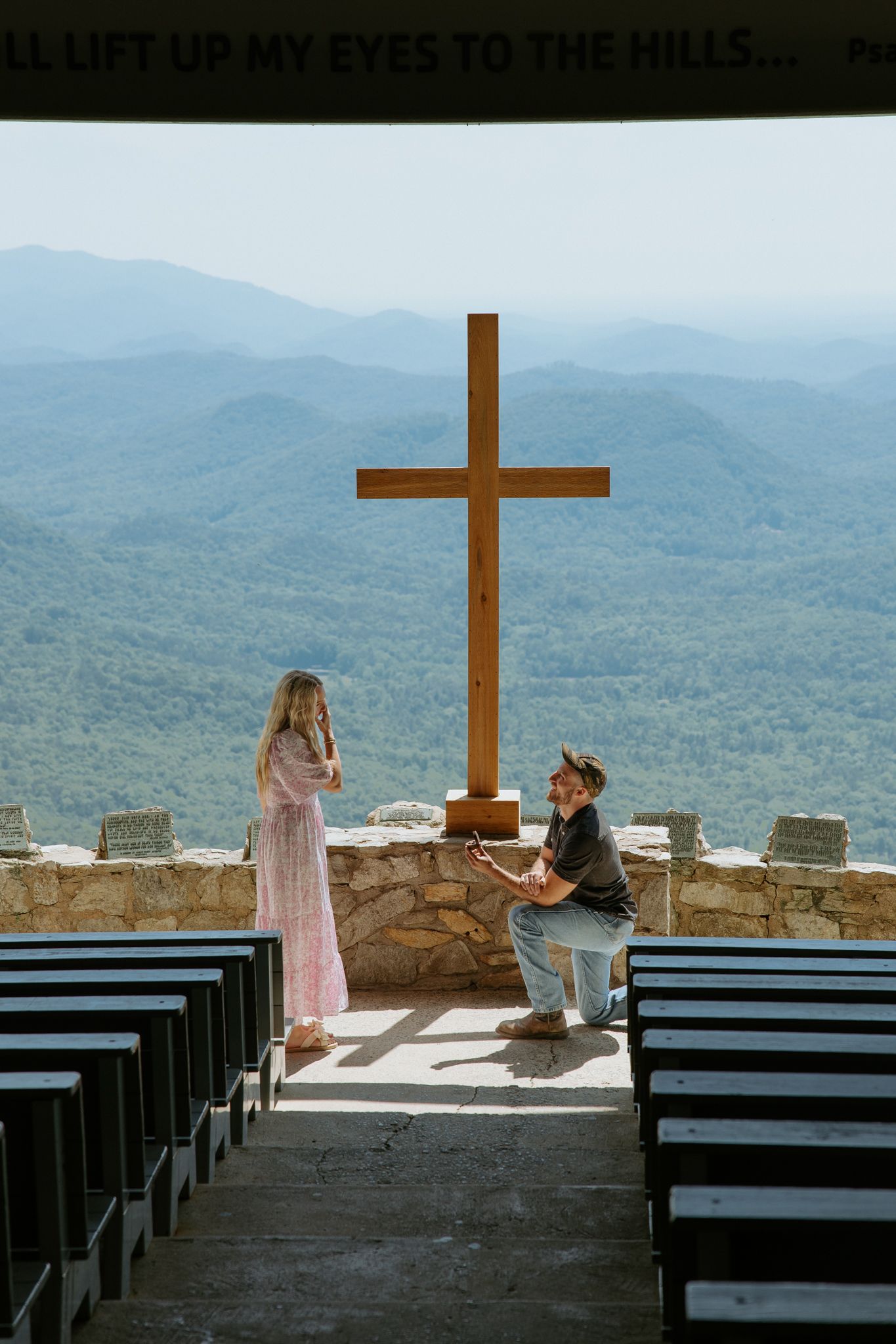 Man proposing to woman in front of large wooden cross at scenic mountain chapel overlook at Pretty Place Chapel.