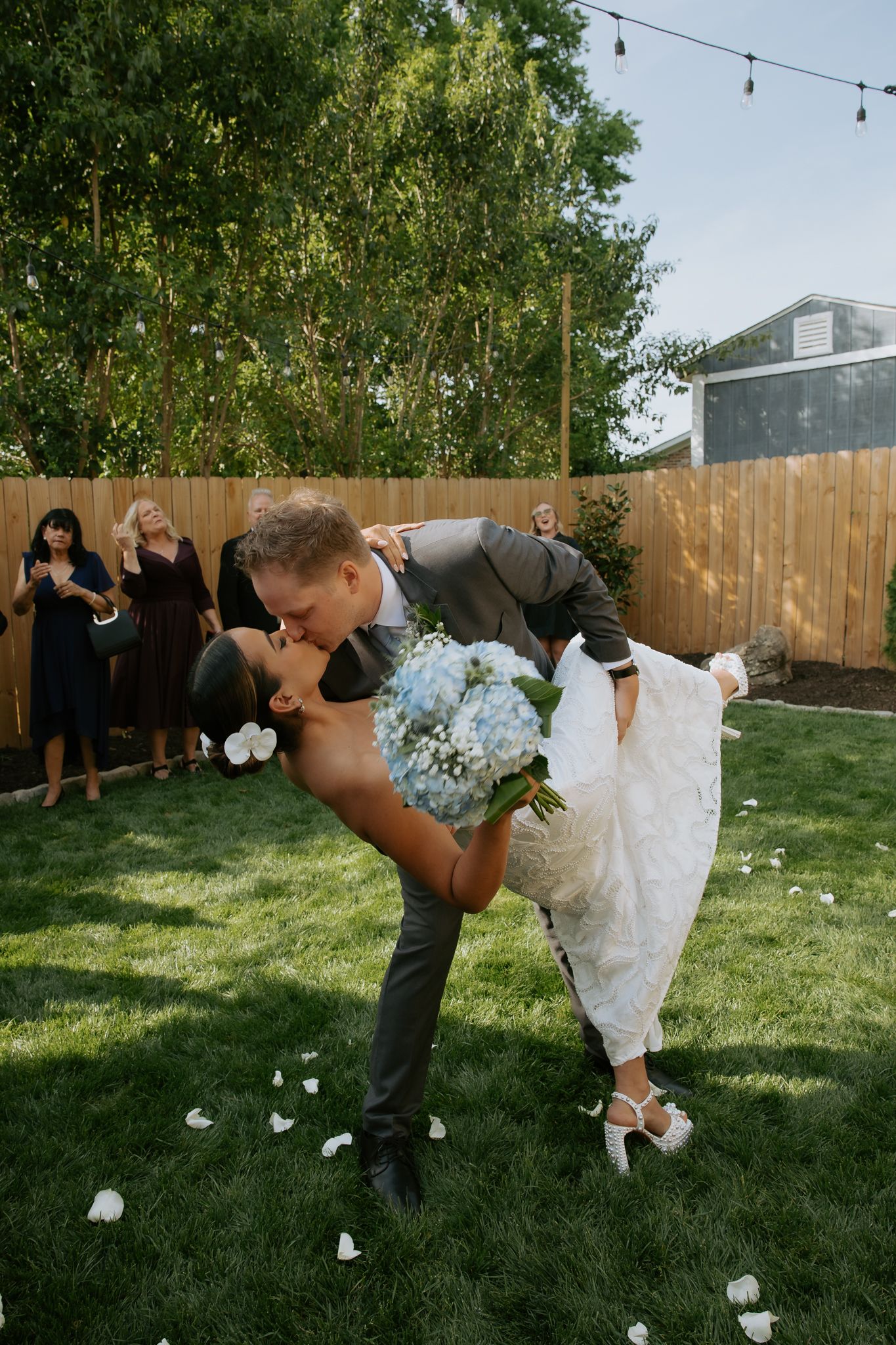 Groom dips bride for a romantic kiss in a backyard wedding, surrounded by flower petals and cheering guests.