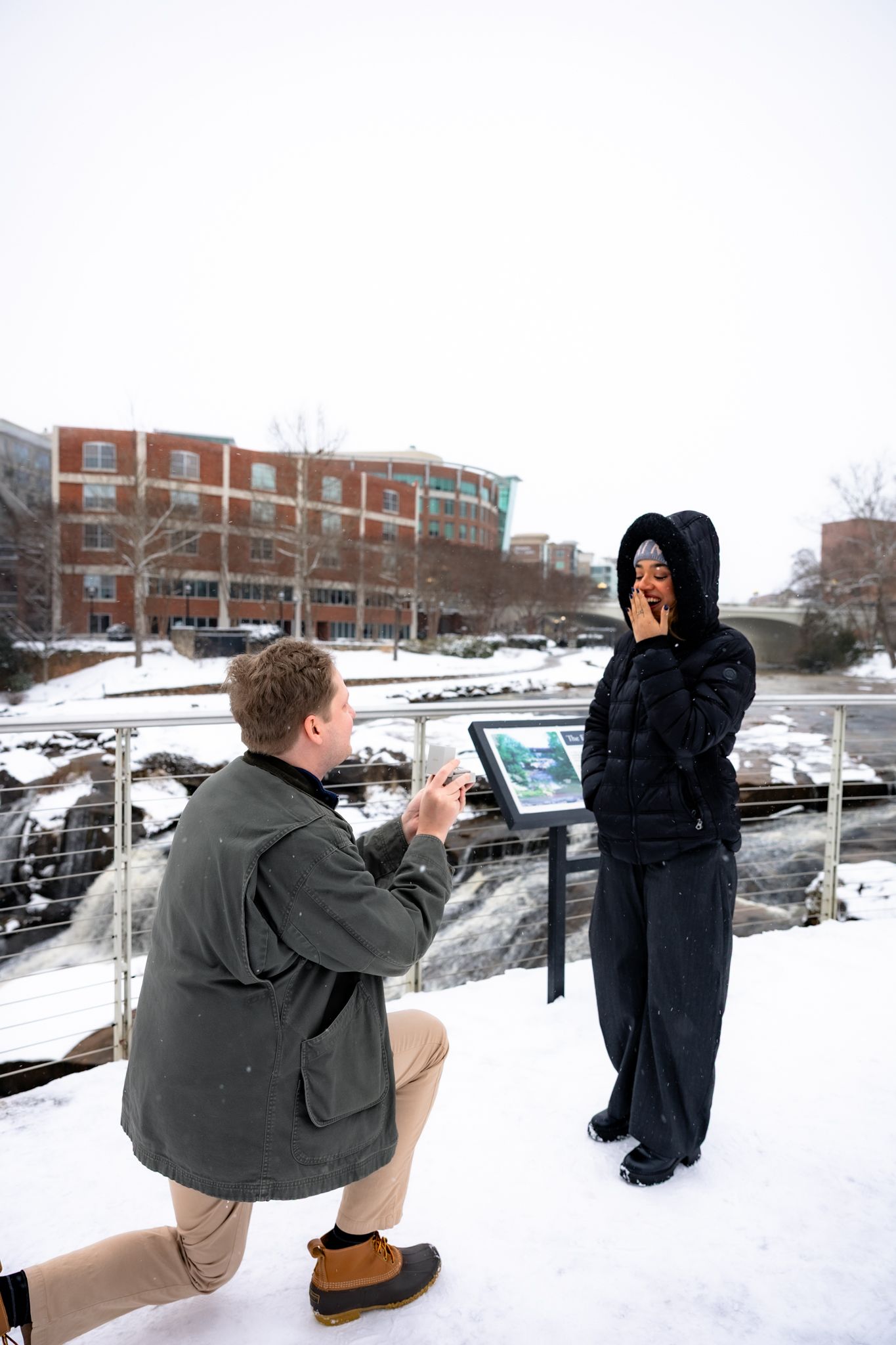 Man proposing on one knee to surprised woman in snowy urban setting near a waterfall at Falls Park in Greenville.