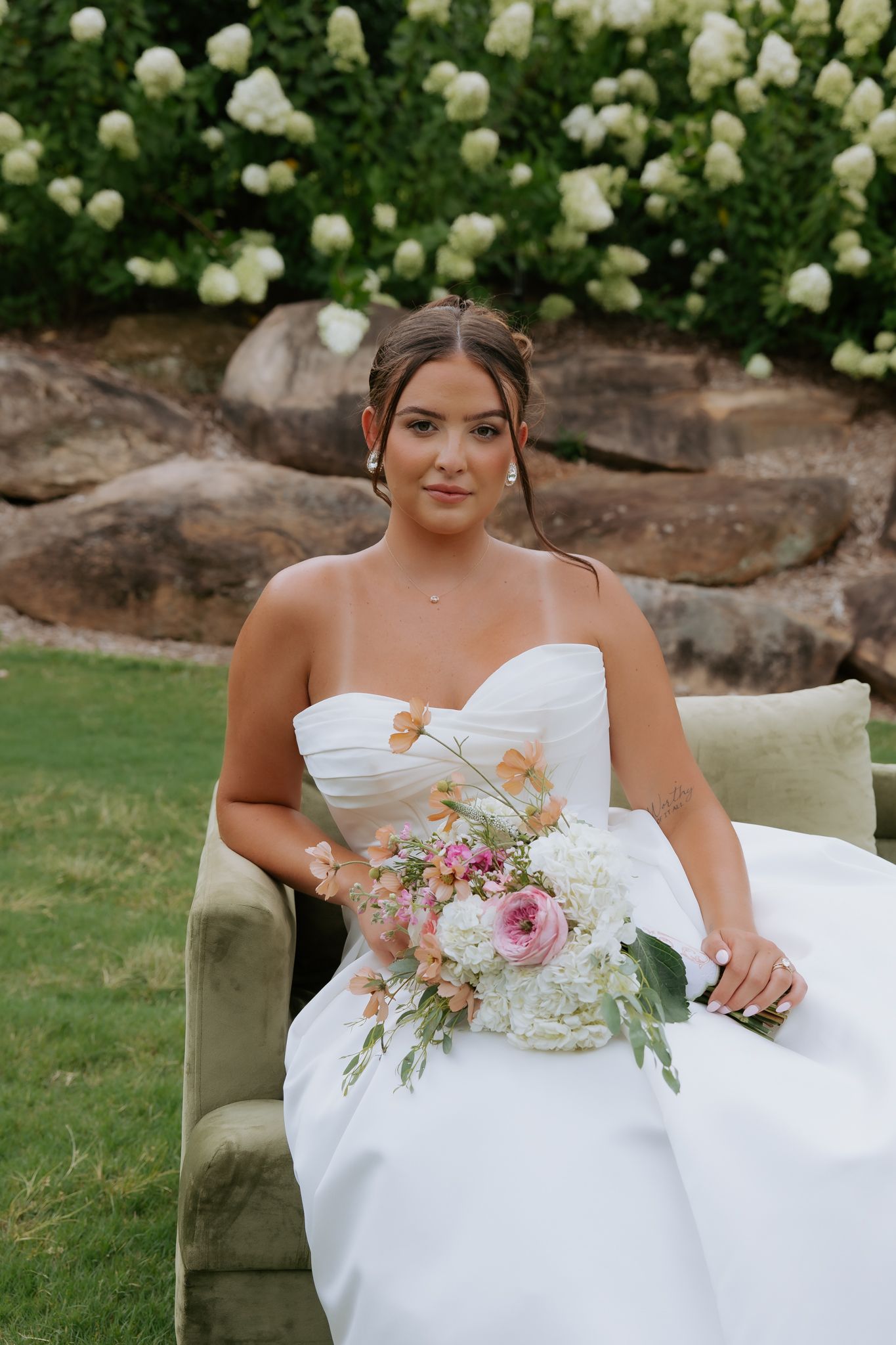 Bride in strapless white gown holds pink and white bouquet seated before white hydrangea bushes at Heyward Manor.