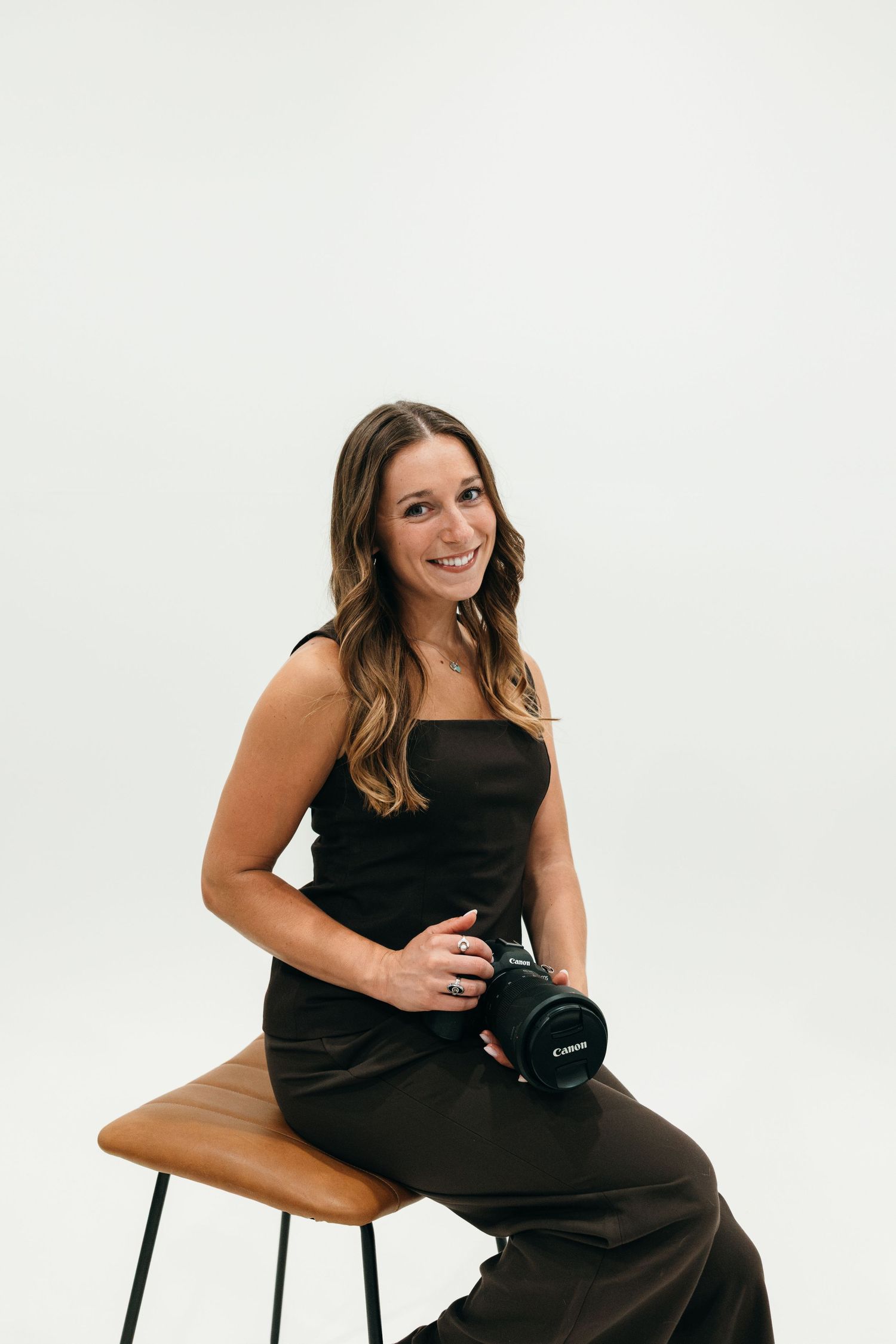 A smiling woman in a black outfit sits on a stool holding a Canon camera as a Greenville Photographer.